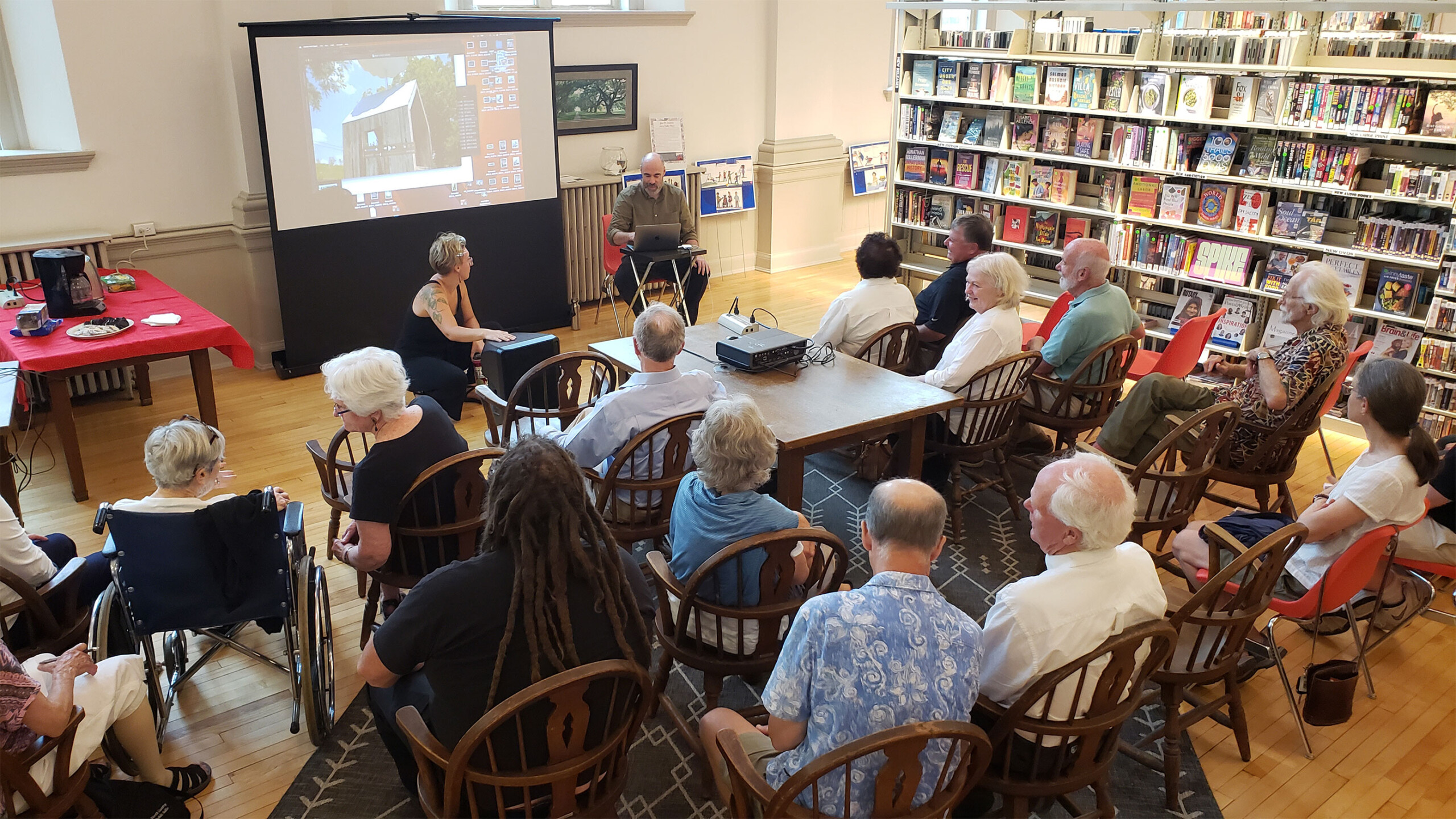 A small group of people gathered in a cozy library setting, seated on wooden chairs arranged in a semi-circle. Books fill the shelves along the walls, creating...