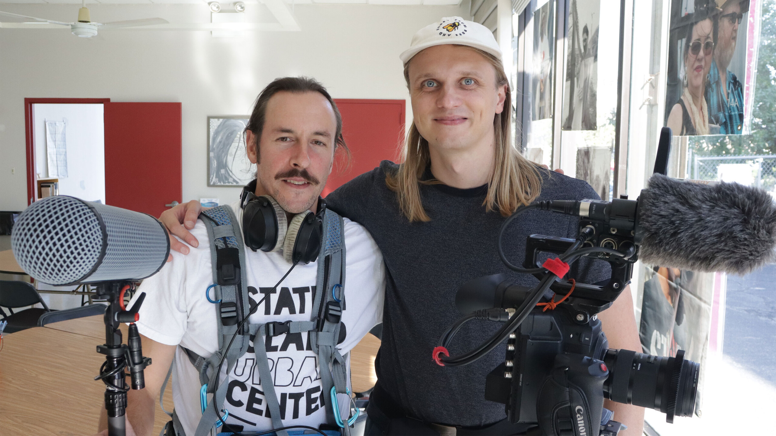 Two individuals smile for the camera in a casual setting. They are equipped with professional audio and video recording gear, including a shotgun microphone and...