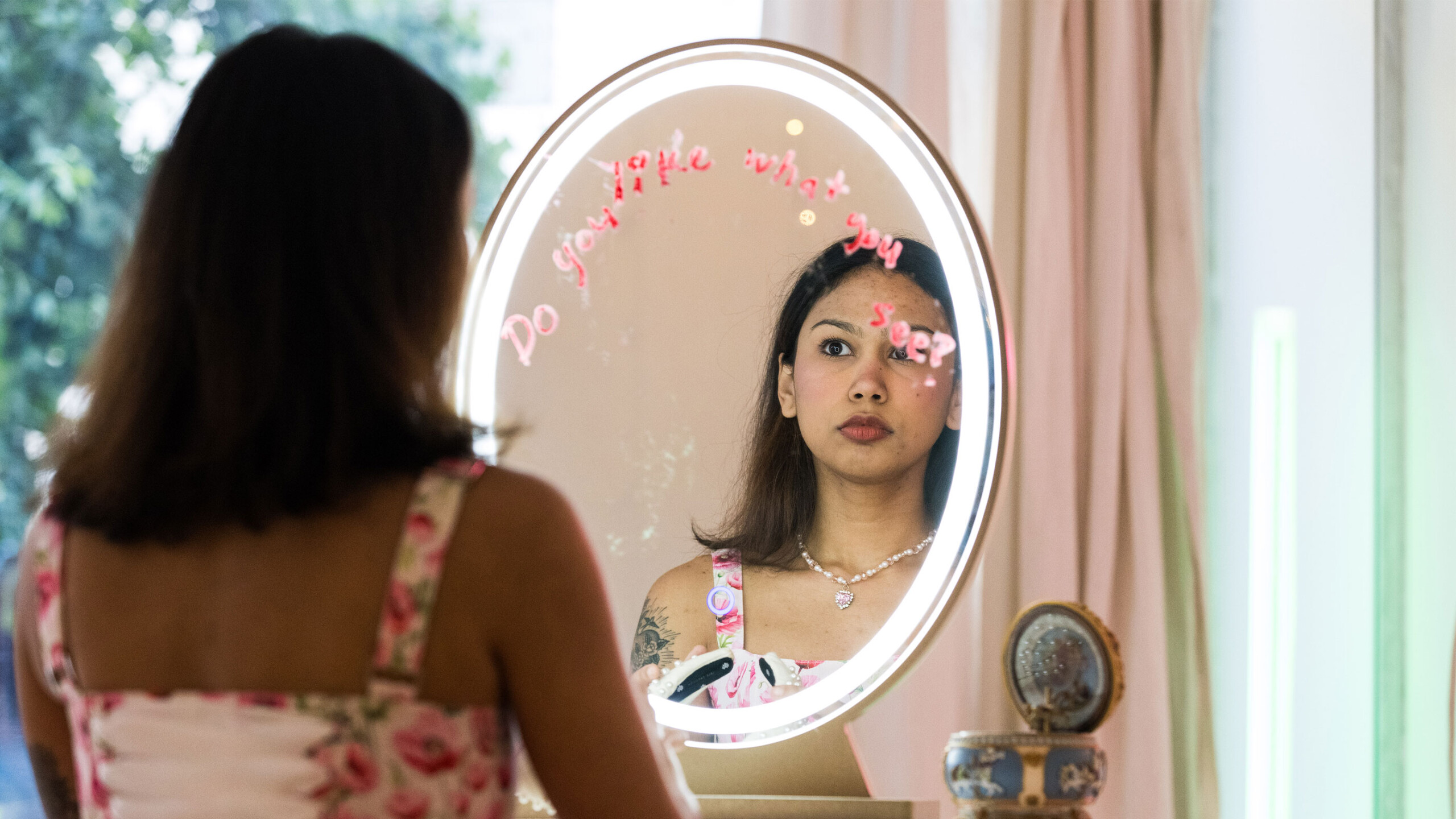 A woman is looking at her reflection in a round, illuminated vanity mirror. The woman in the reflection is light-skinned, with brown hair styled in a medium length...