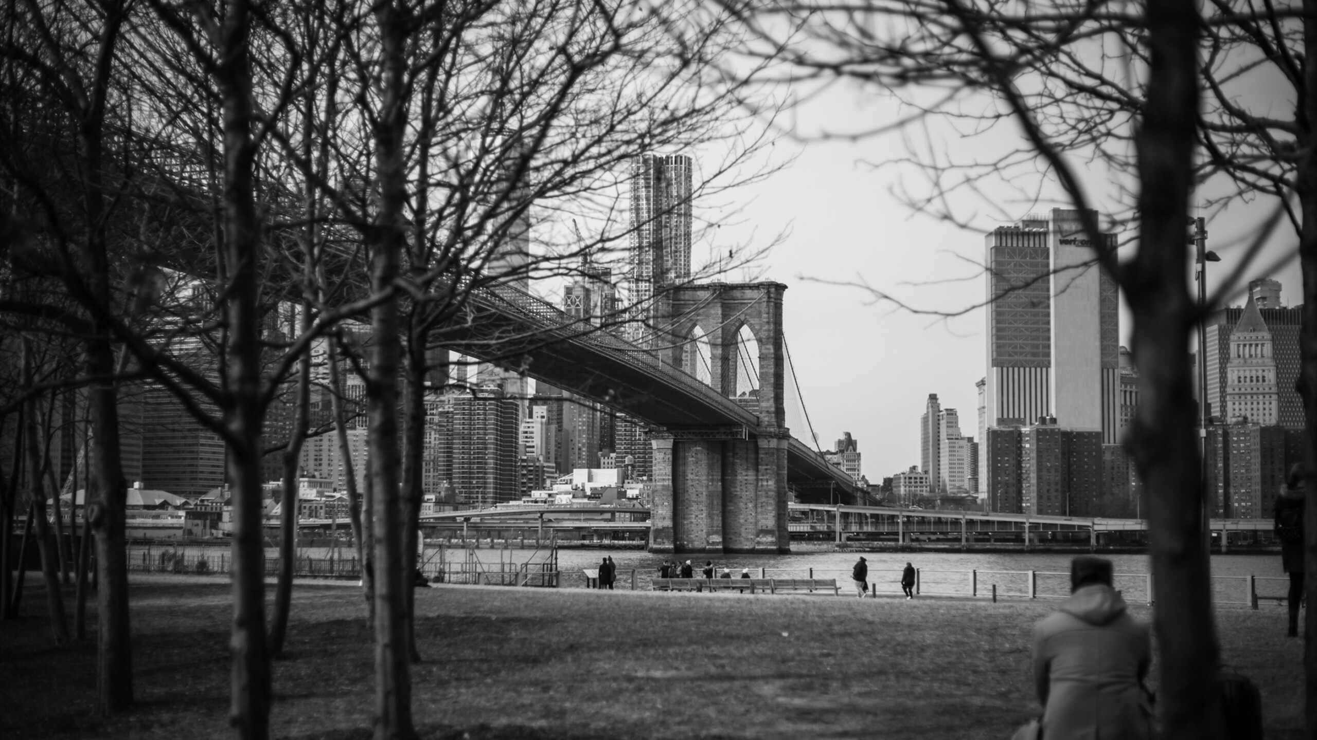 A monochrome image featuring the iconic Brooklyn Bridge stretching across the East River in New York City. The bridge stands prominently in the foreground, framed...