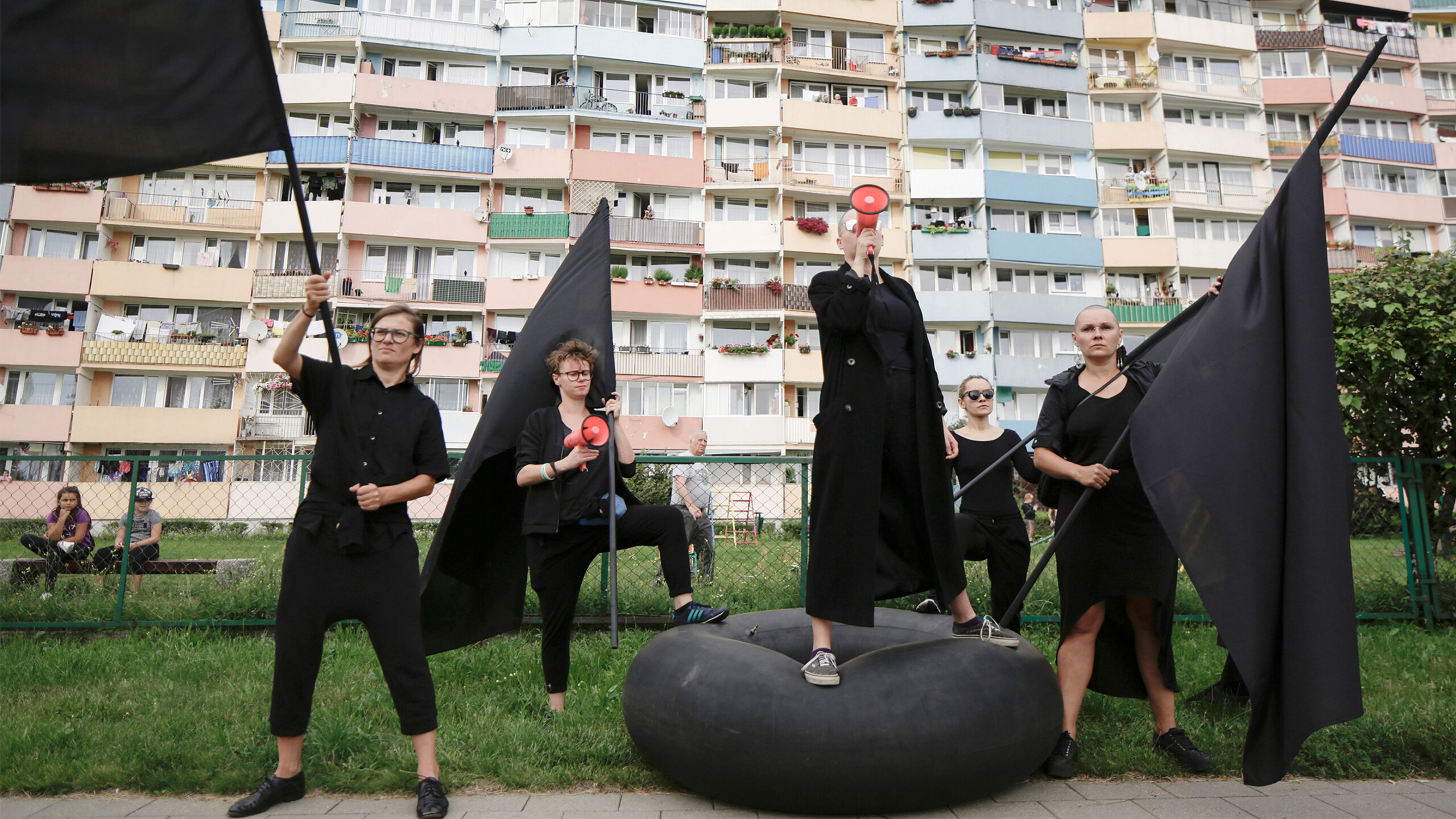 A group of individuals dressed in black clothing stand confidently holding large black flags. They are positioned on a grassy area in front of a colorful apartment...