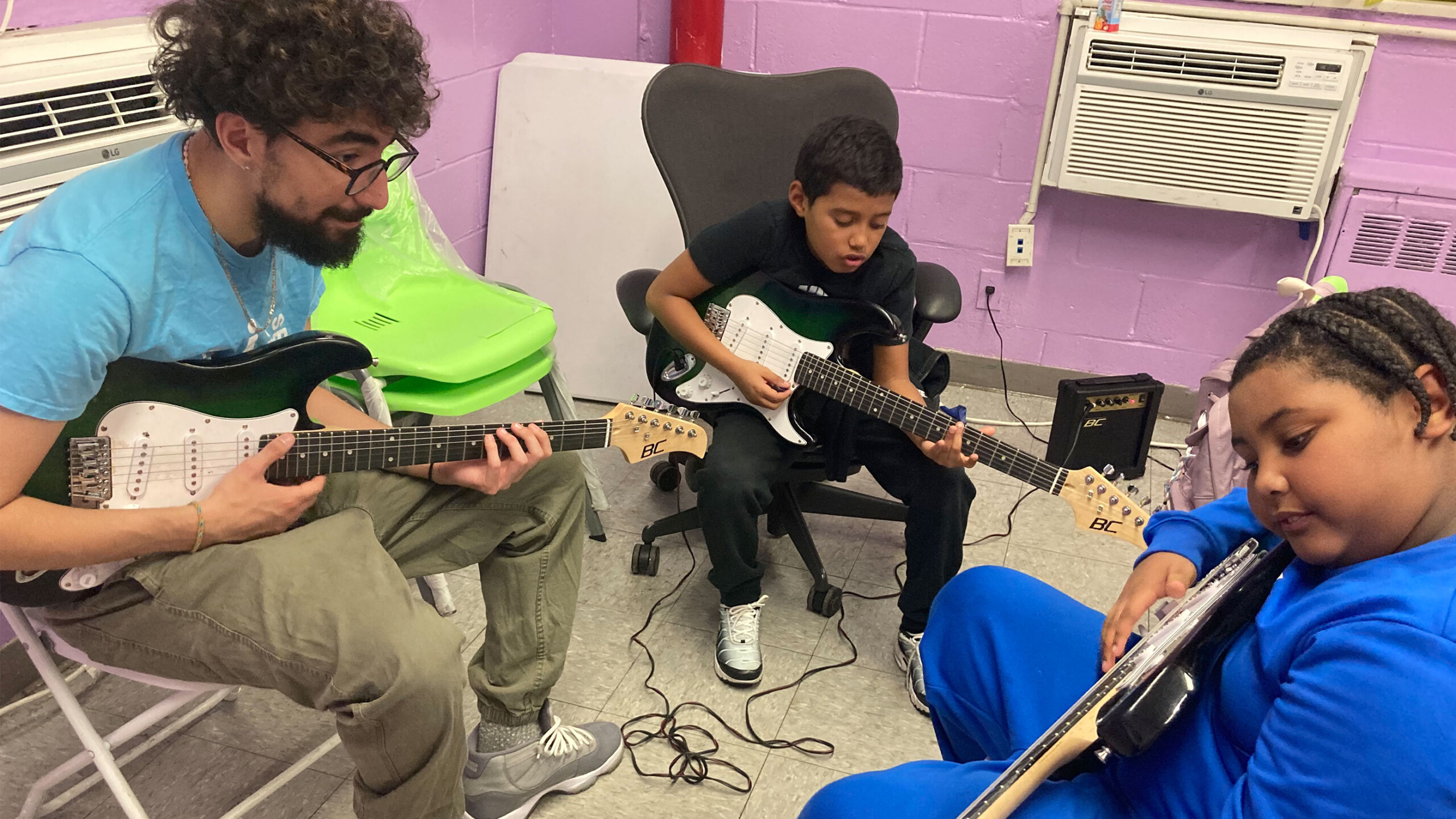Three individuals are engaged in a guitar practice session. Two children, a boy and a girl, are holding electric guitars, focused on their playing. The boy is seated...