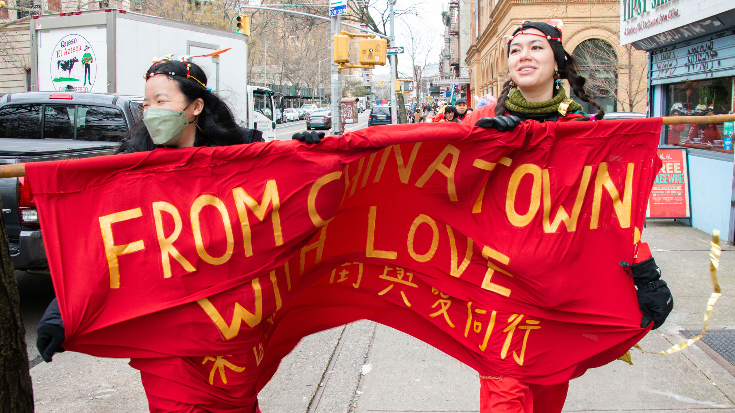 Two individuals walking down a city street holding a vibrant red banner with the message 'FROM CHINATOWN WITH LOVE' written in bold, yellow letters. Below the English...