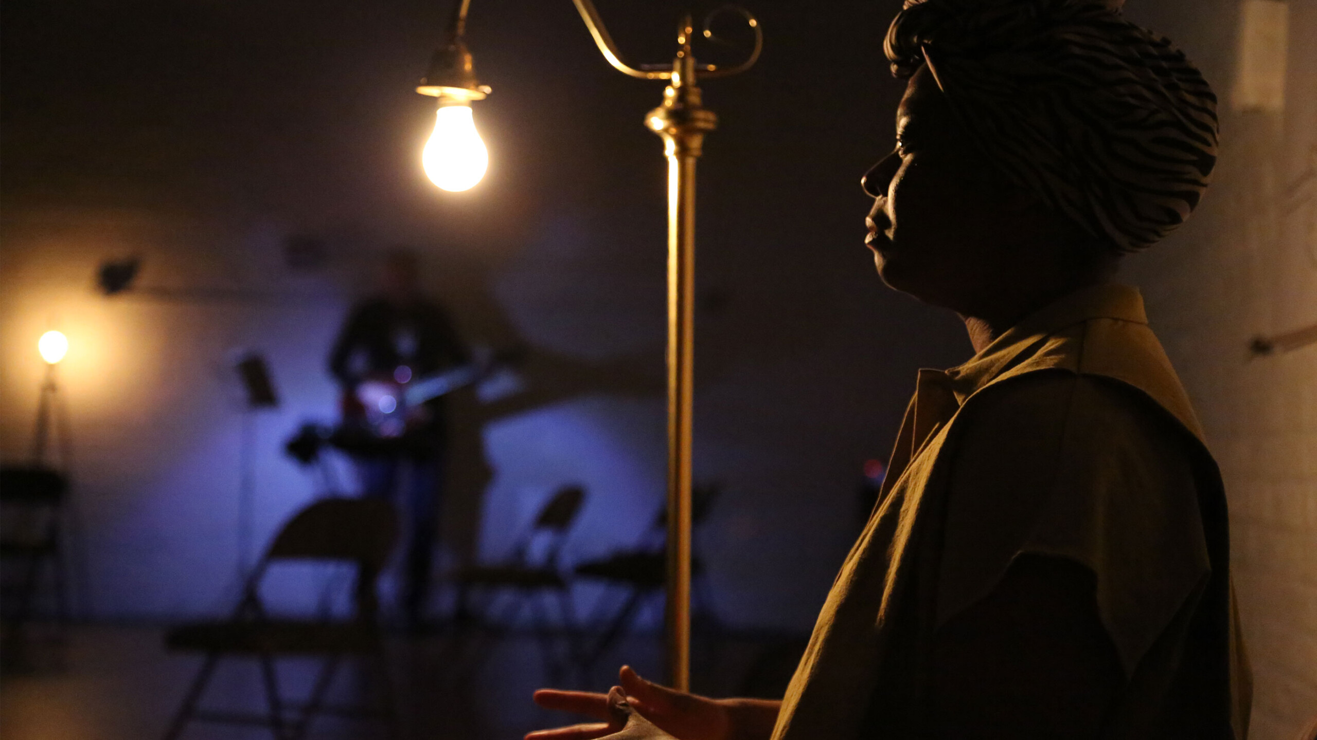 A dimly lit room features a dramatic scene illuminated by an overhead lightbulb, casting shadows and highlighting textures. In the foreground, a person is silhouetted,...