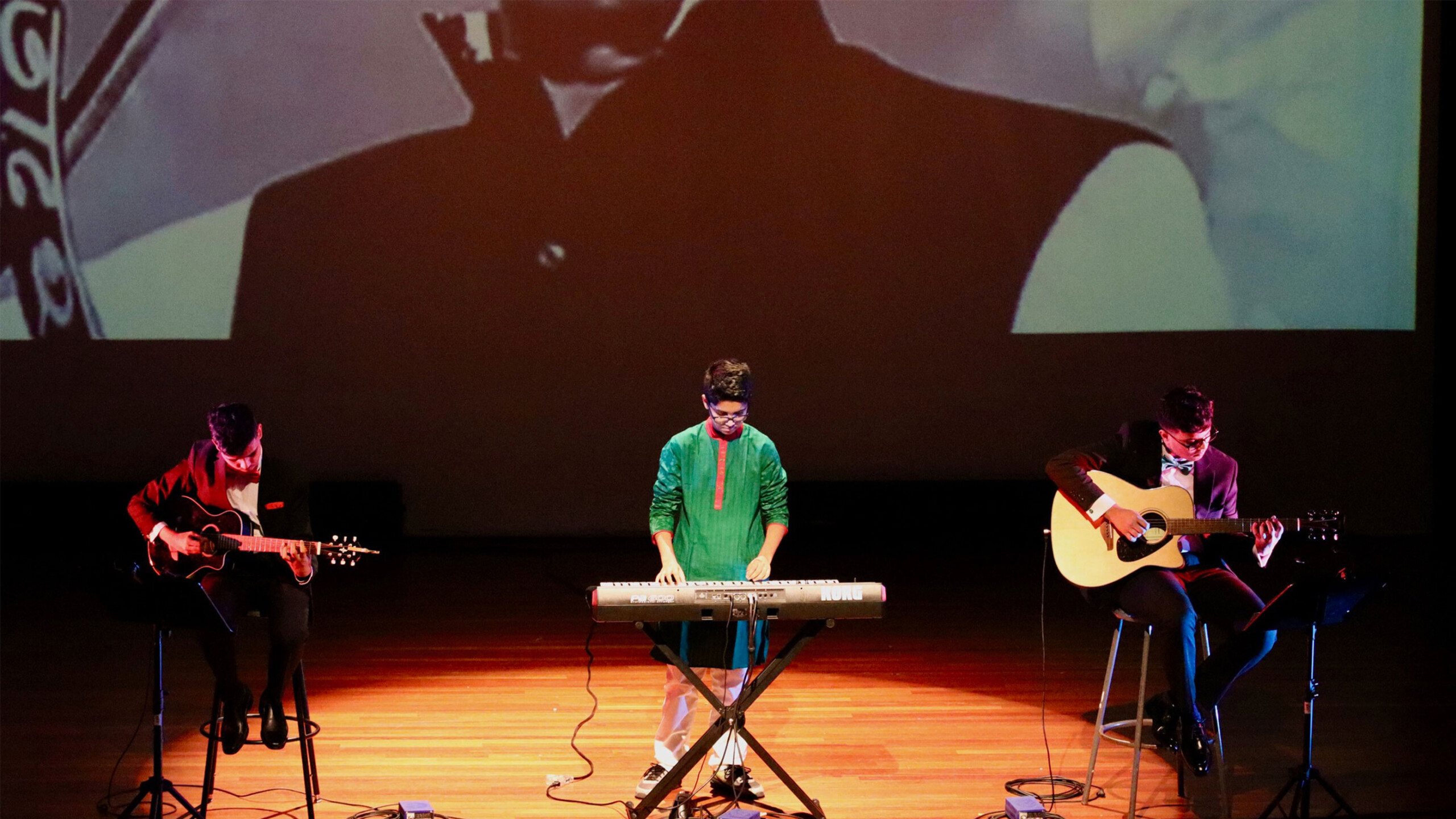 Three musicians playing instruments on stage with a large projection screen in the background.