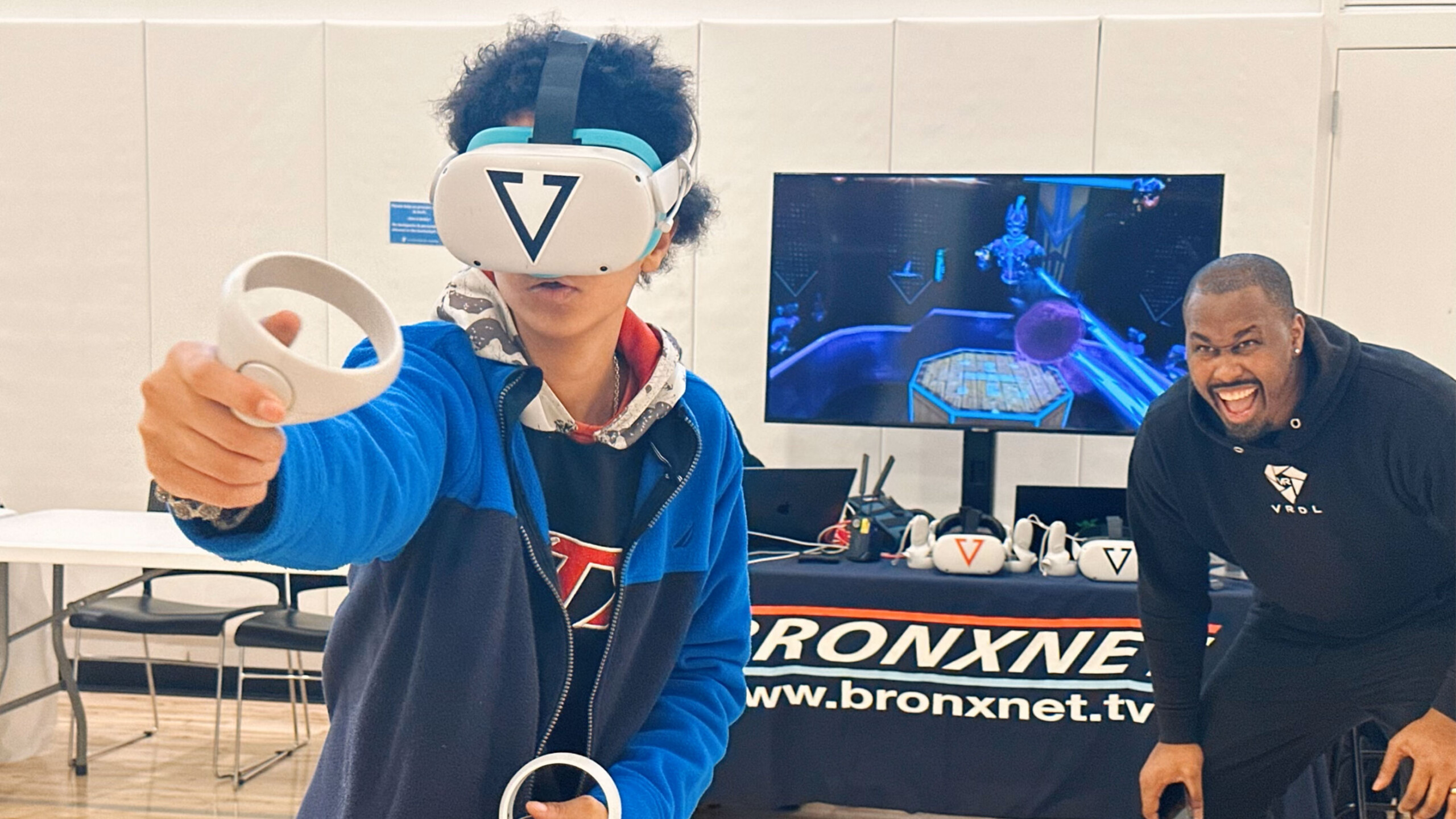 A person wearing a VR headset and holding controllers engages in a virtual reality experience, fully immersed and actively participating. Behind them, a large monitor...