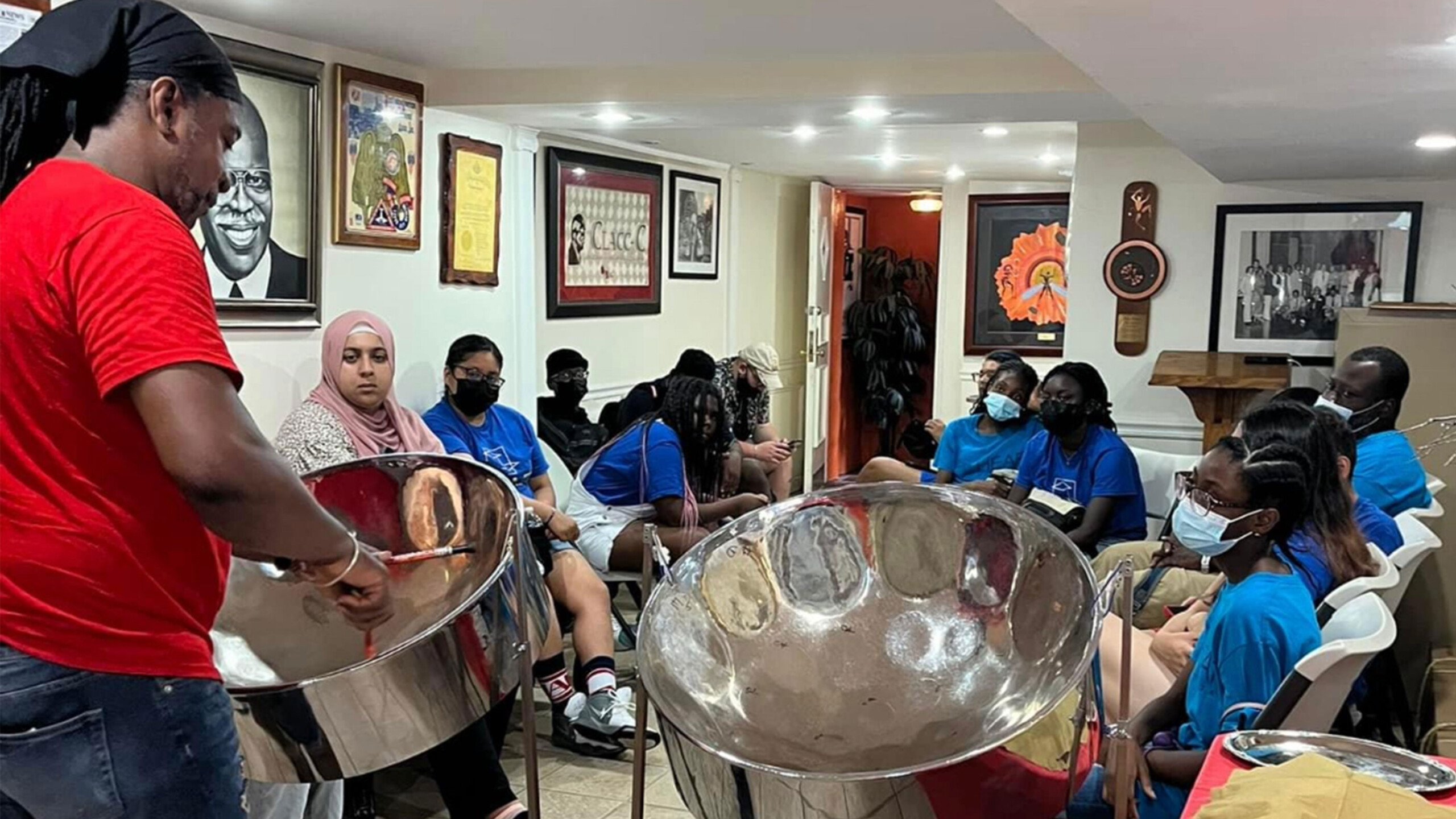 A group of people is gathered in a room, engaged in a music session focused on steel pan drums. In the foreground, a person wearing a red shirt plays the steel...