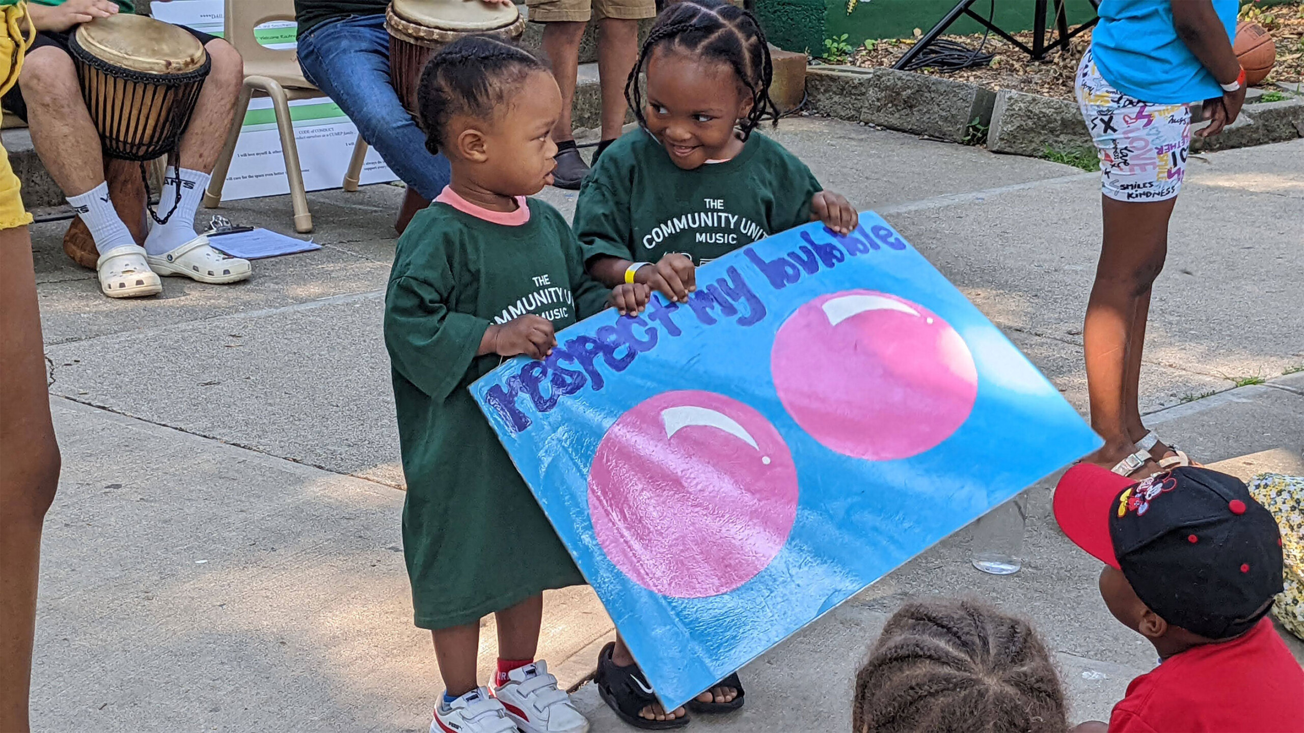 Two young children stand on a concrete surface, both dressed in oversize green t-shirts. They are holding a large, colorful poster that reads 'Respect My Bubble'...