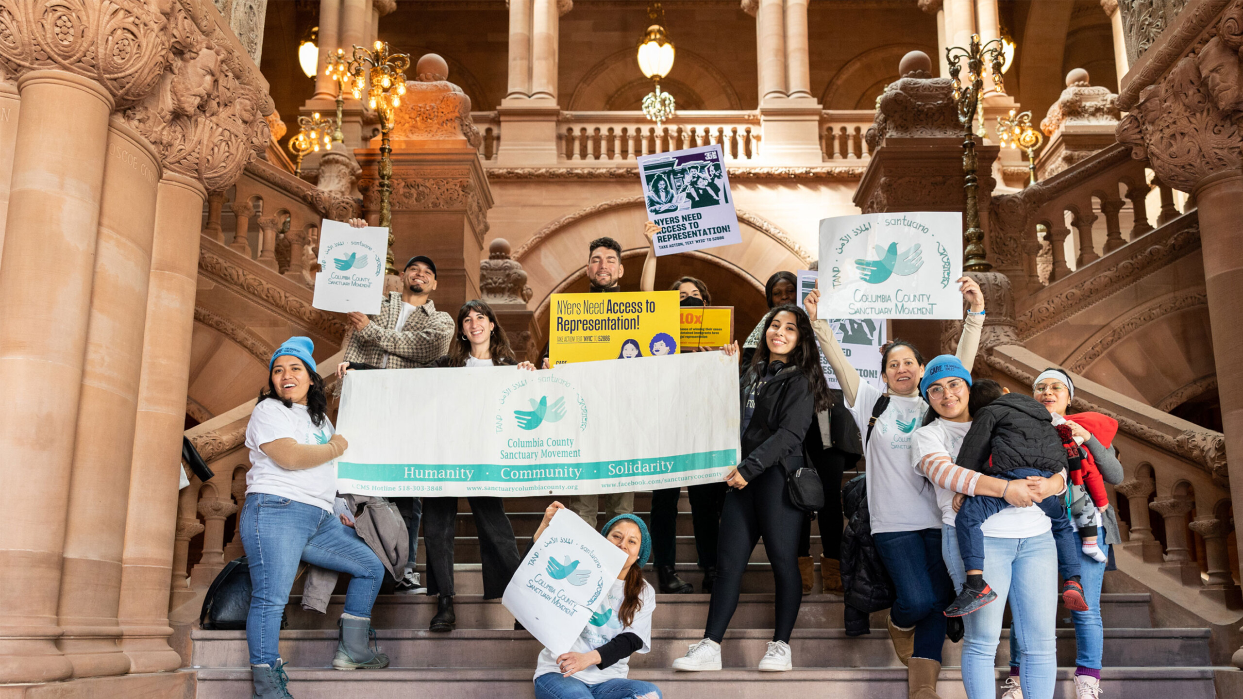 A diverse group of people standing on a grand, ornately decorated staircase in what appears to be a government building or significant public venue. The architecture...