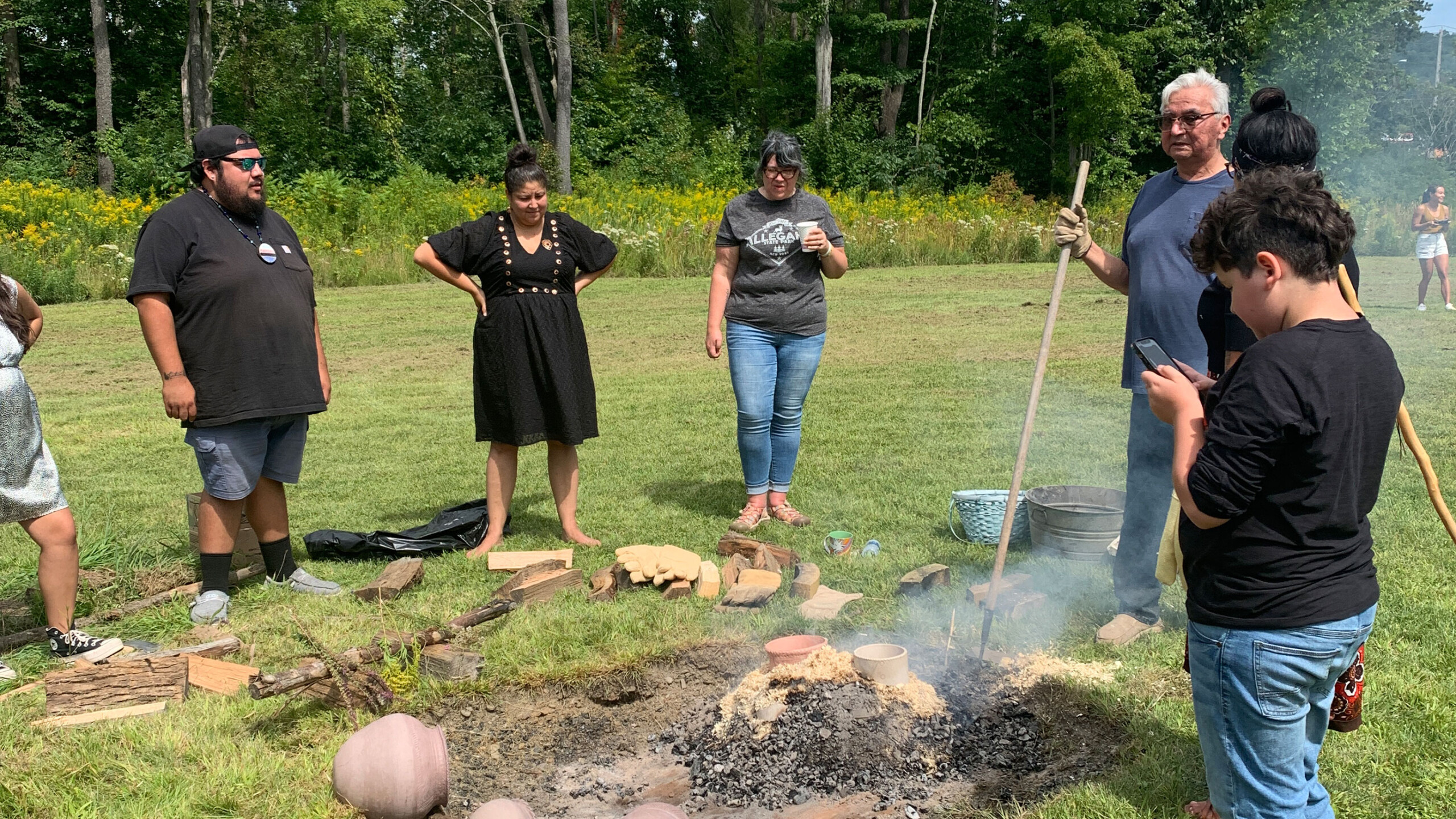 A group of people standing outdoors on a grassy field, surrounding a small open fire pit. Smoke rises from the fire where a collection of pottery and clay objects...