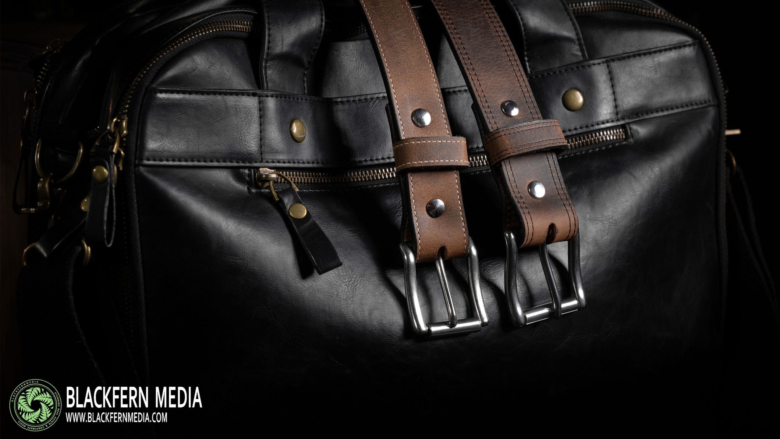 A close-up view of a sophisticated black leather bag with elegant design elements. Two brown leather straps with silver-tone buckles are prominently featured, adding...