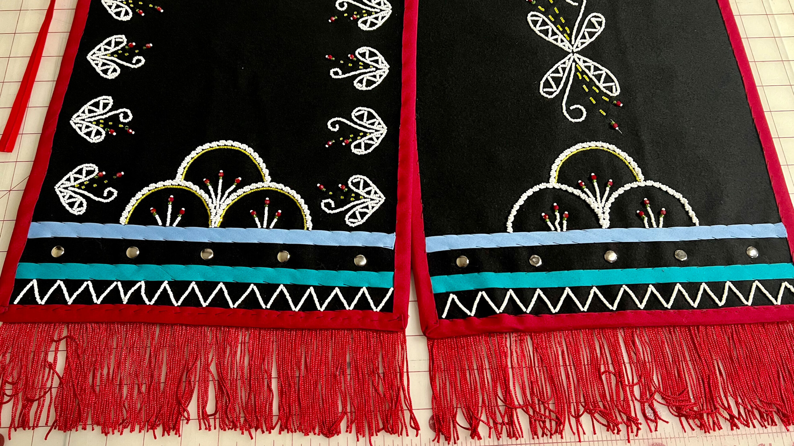 Two pieces of black fabric adorned with intricate white embroidery designs featuring symmetrical floral and nature-inspired patterns. The fabric is bordered by...