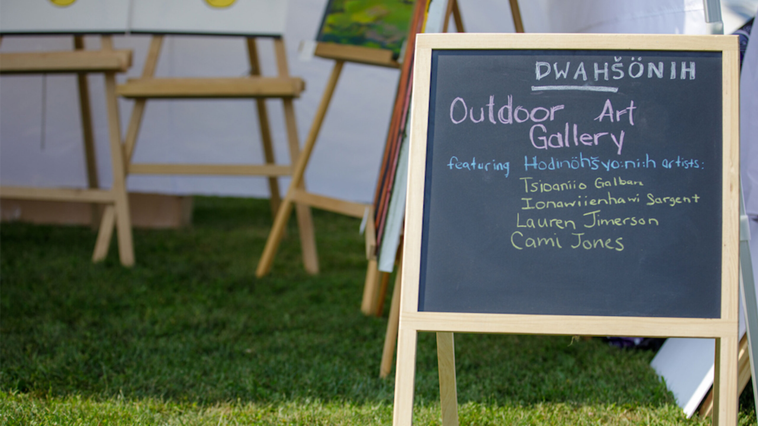 A wooden easel holds a chalkboard sign on a patch of green grass. The sign reads 'Dwa’shonih Outdoor Art Gallery' and lists several artists: Tsonias Galban, Ionawrienthanhan...