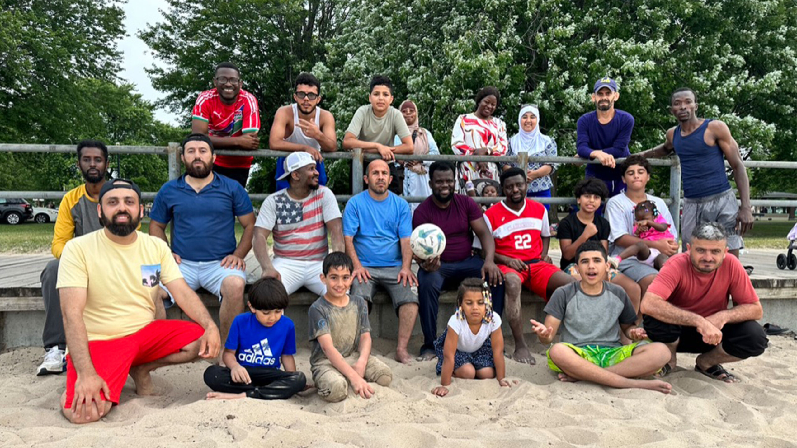 A diverse group of people gathered on a sandy beach, including men, women, and children, most of whom are seated on the sand or standing on a wooden walkway. They...