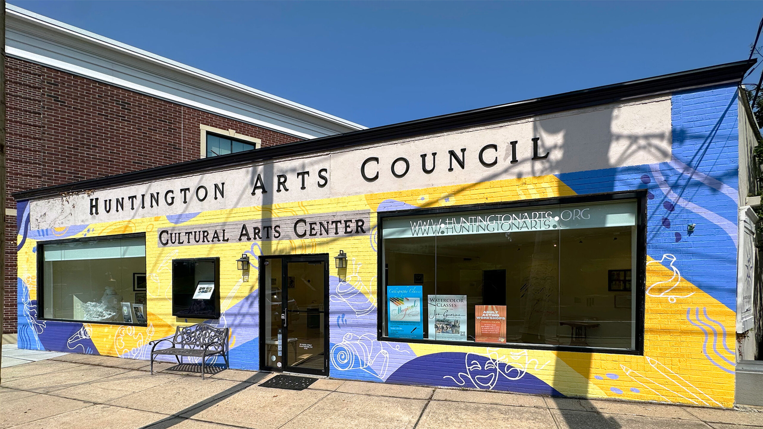 A vibrant facade of the Huntington Arts Council building, featuring a colorful mural with abstract patterns in yellow, blue, and purple hues. The words 'Huntington...