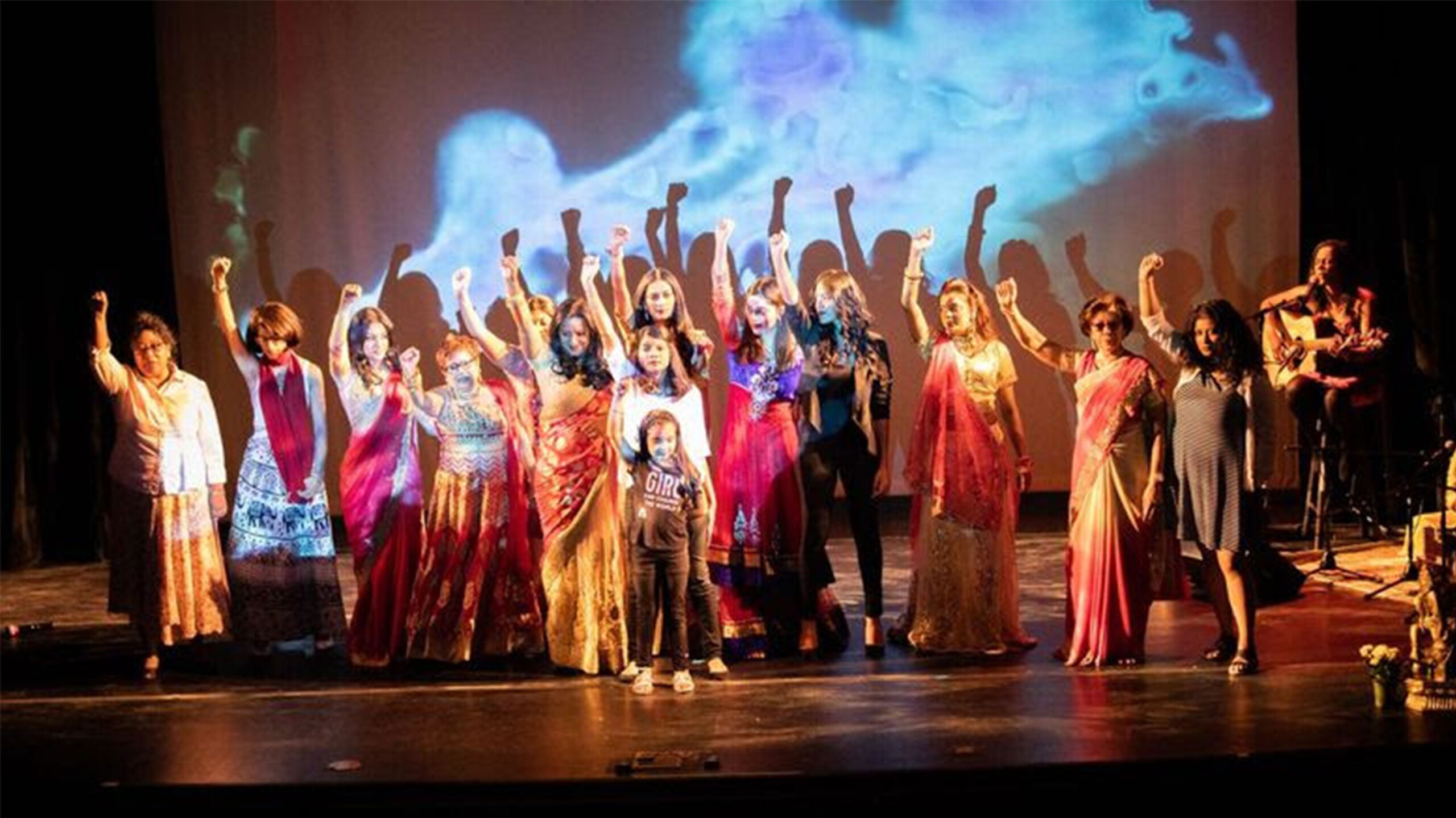 A group of diverse women and a child stand on a stage, holding their fists in the air in a gesture of empowerment and solidarity. They are dressed in a variety...