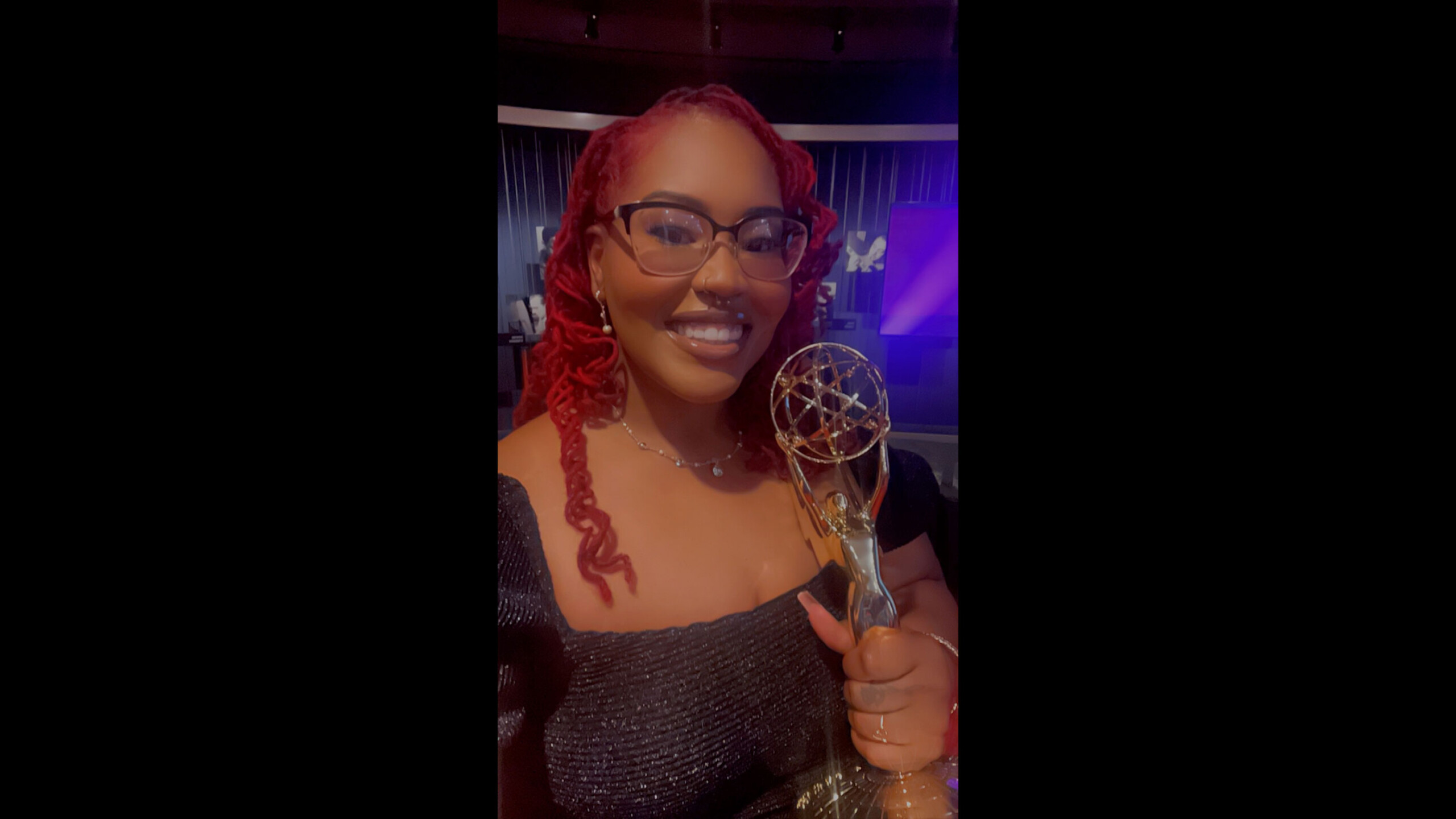 A woman with vibrant red hair and glasses smiles broadly while holding a prestigious award. She wears a shimmering dark dress with off-the-shoulder sleeves.