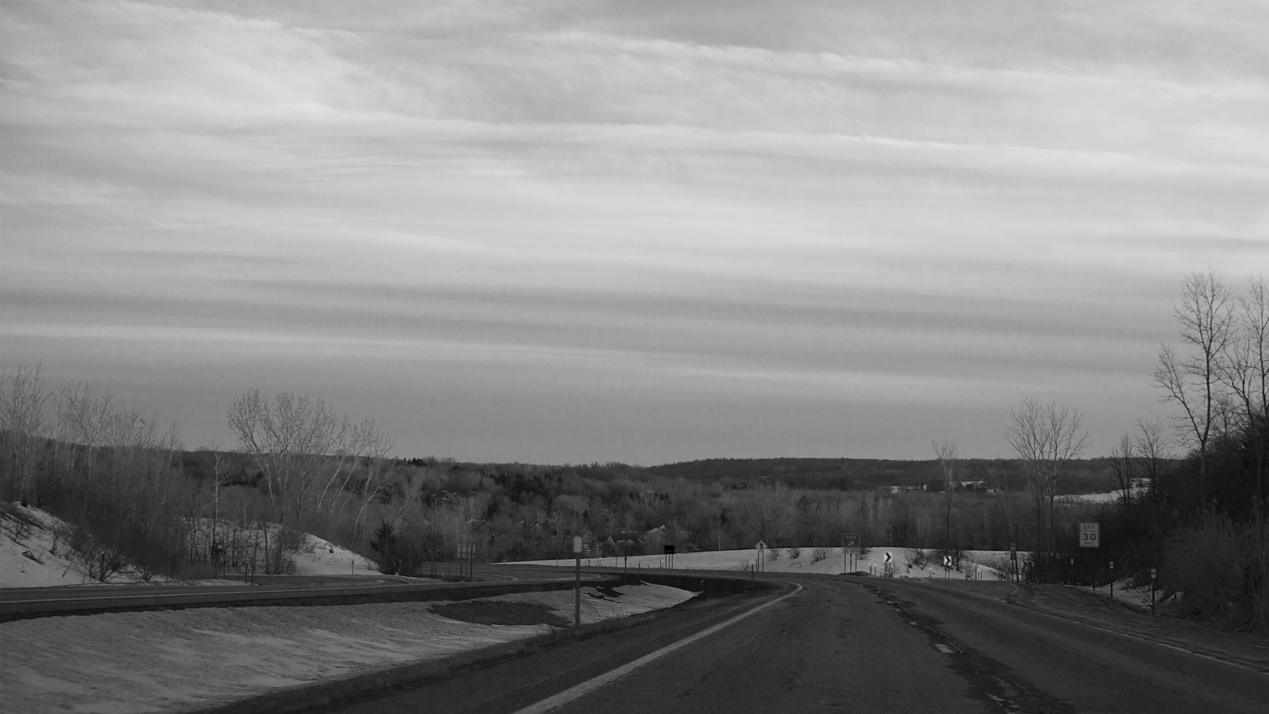 A deserted road stretches into the distance, flanked by snow-covered shoulders and barren trees. The landscape is monochrome, with bare trees lining the road, their...