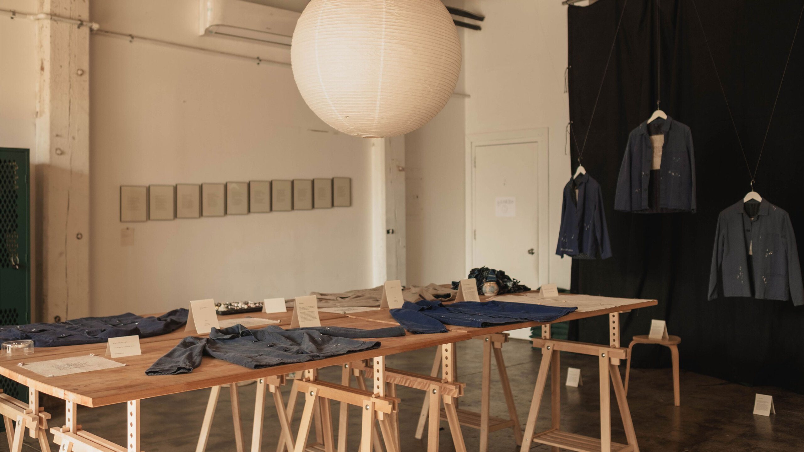 A spacious exhibition room featuring a large, spherical paper lantern hanging from the ceiling, casting soft light over the space. A series of wooden trestle tables...