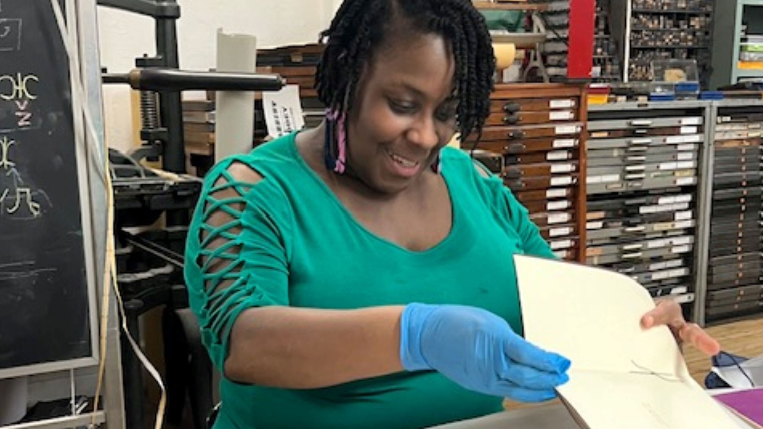 A woman with braided hair, wearing a green top with cut-out sleeves, is bookbinding in a printshop. She has a gentle smile and is engaged in her work, holding a...