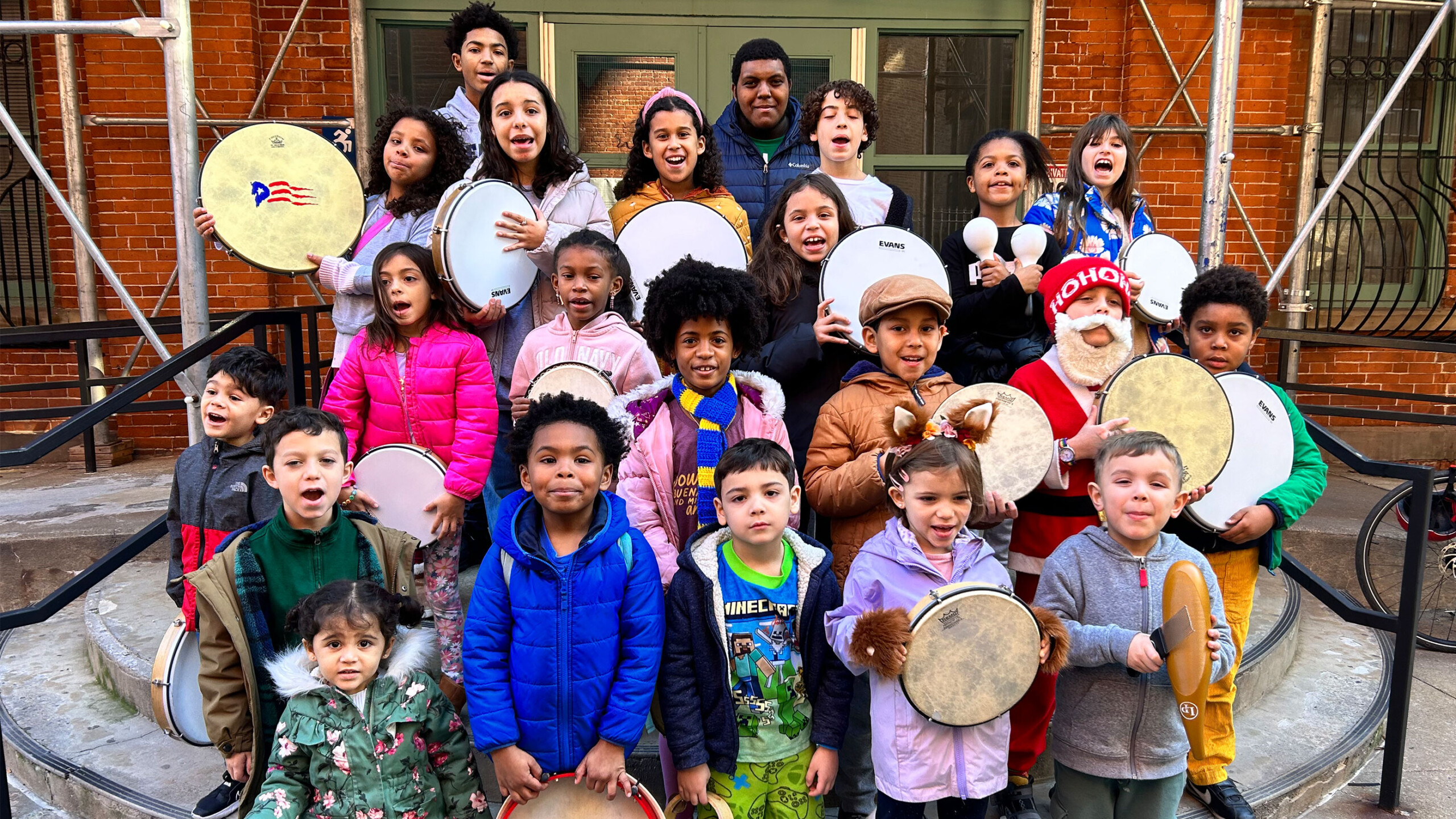A group of children of various ages gather on the steps of a building, each holding a tambourine. The children are diverse, dressed in colorful winter clothing,...