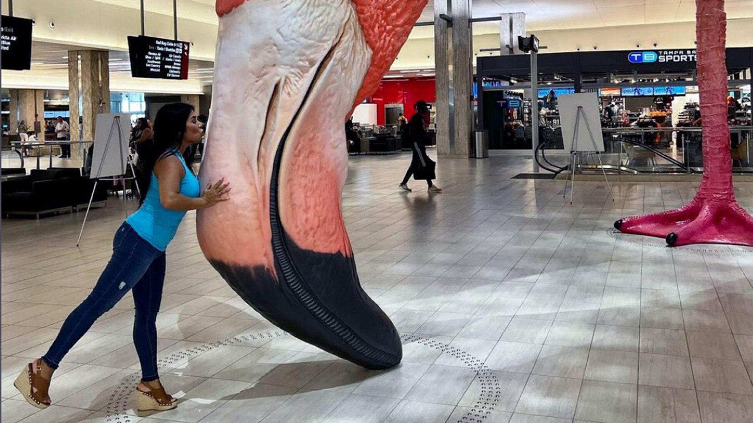 A woman is playfully pressing against a massive, realistic sculpture of a flamingo's leg inside a spacious, modern indoor area. The leg is predominantly pink with...