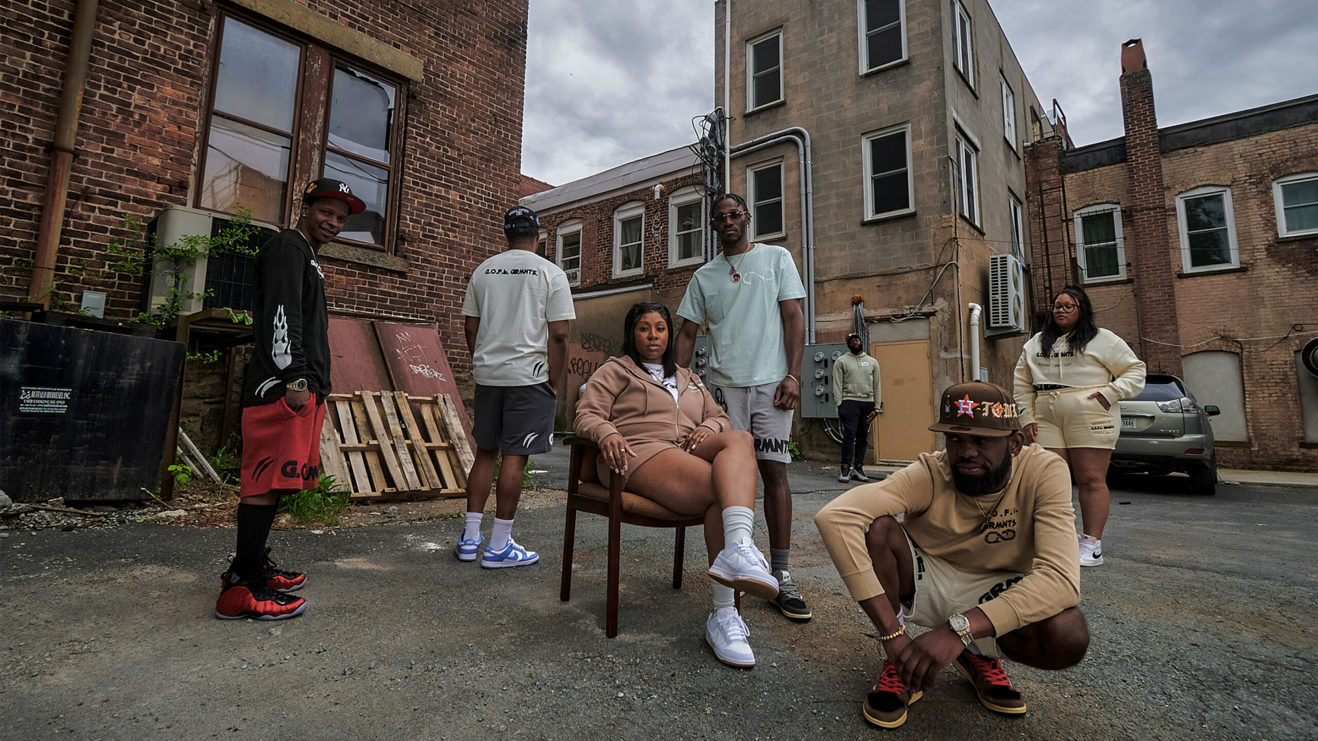 A group of six people poses in a gritty urban alleyway, surrounded by brick buildings and a cloudy sky. The individuals wear trendy streetwear, including hoodies,...