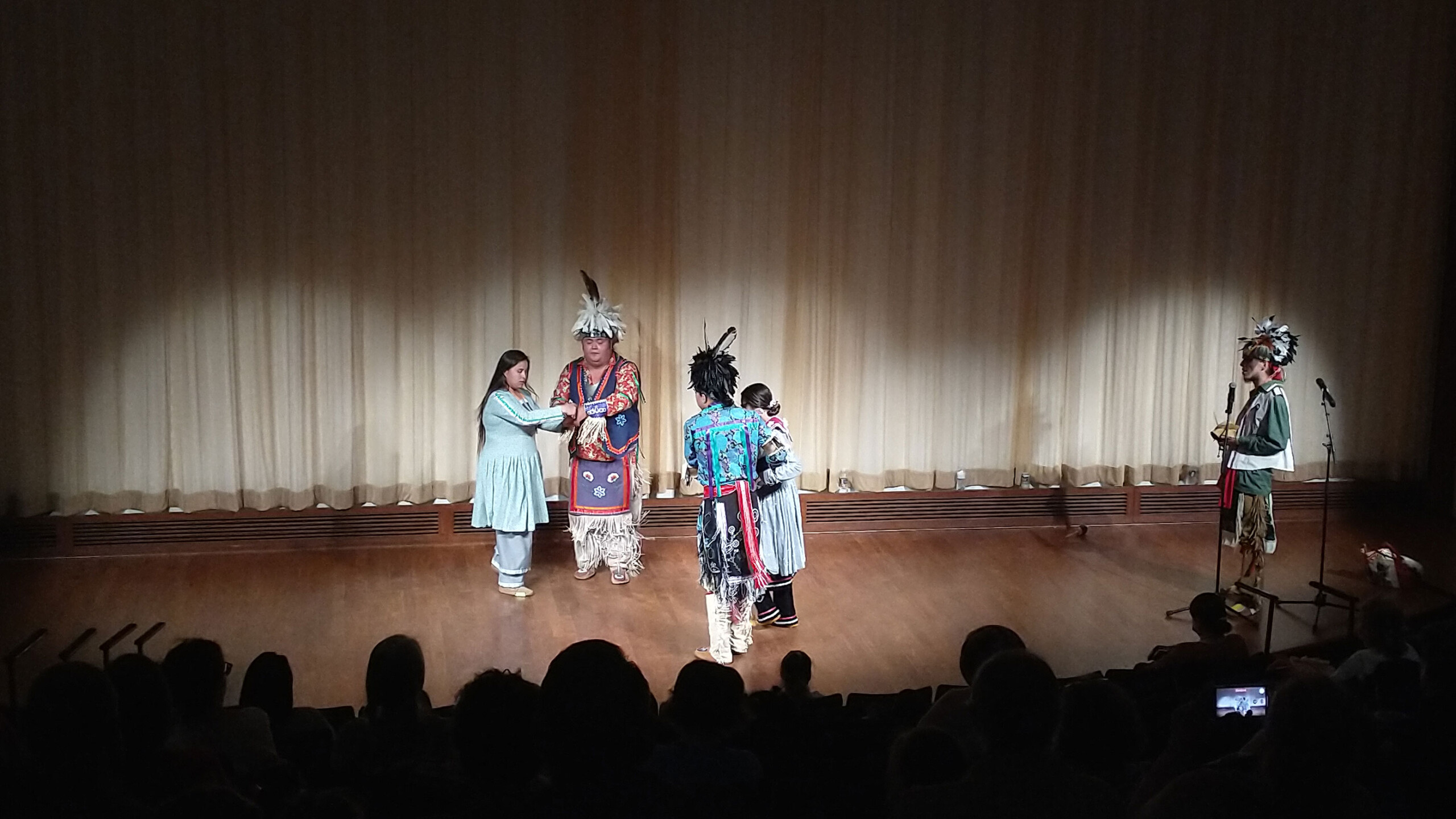 A group of performers wearing traditional indigenous attire are gathered on a dimly lit stage. The scene features intricate costumes decorated with vibrant colors...