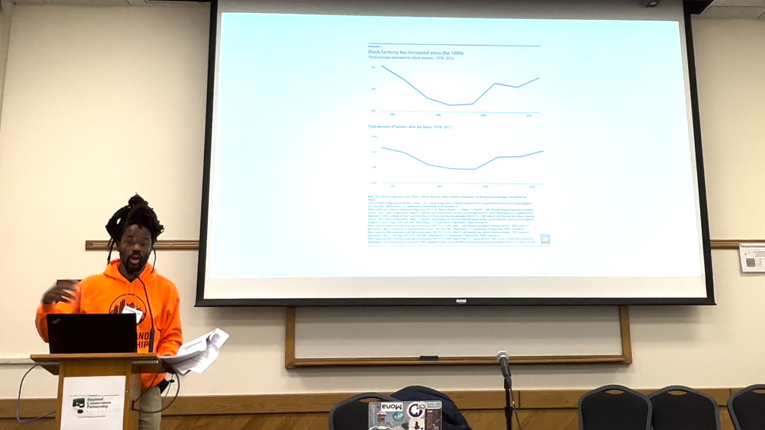 A person is sitting at a desk in front of a projector screen. The setting appears to be an educational environment, likely during a presentation or lecture. The...