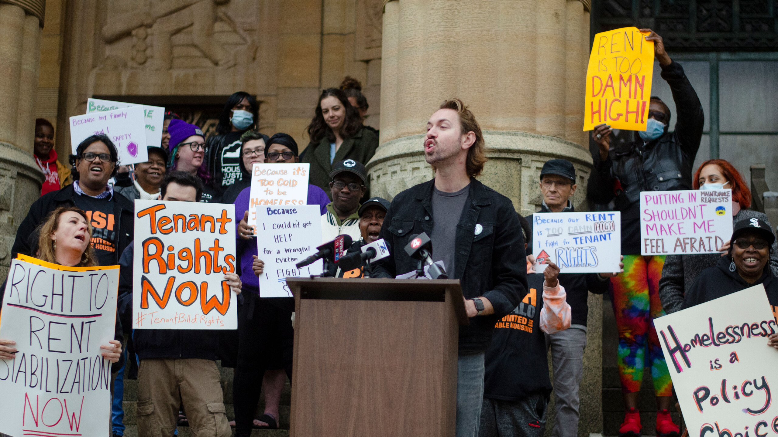 A group of protesters gathered on the steps of a building, advocating for tenant rights and rent stabilization. The central figure, a person speaking passionately...