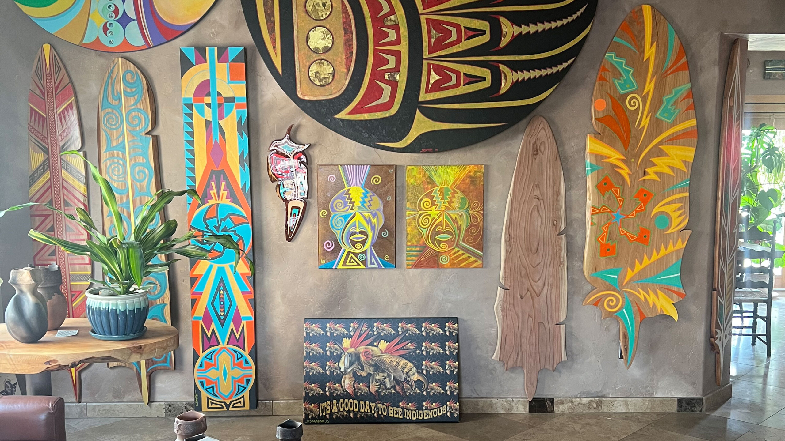 A dynamic art display features surfboard-shaped pieces and colorful murals on a wall. Various elements include a large circular tribal design at the top and elongated...