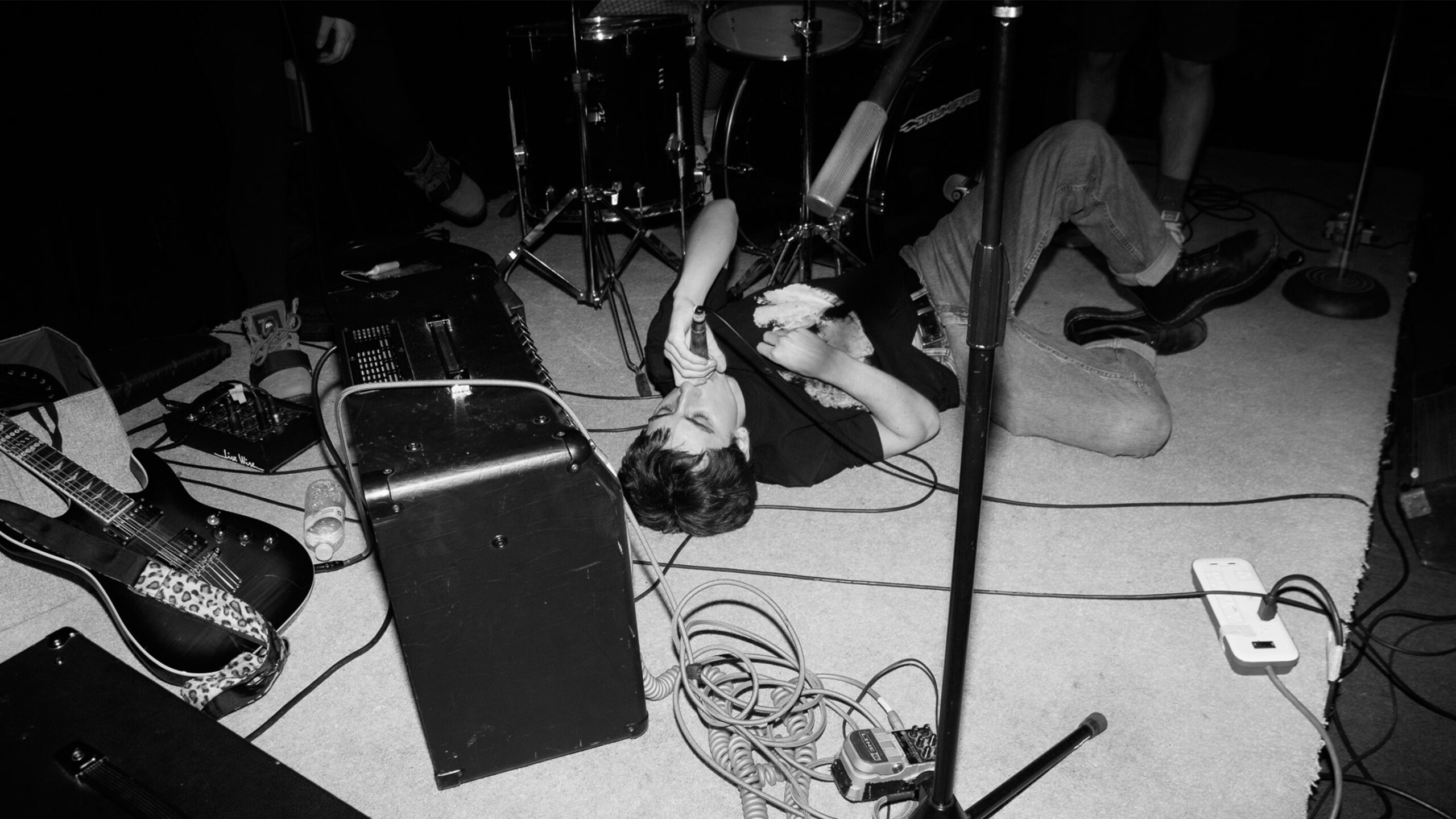 A performer is lying on the floor amidst a chaotic scene of musical equipment during a live performance. The setting is a small stage with visible drum kit components...