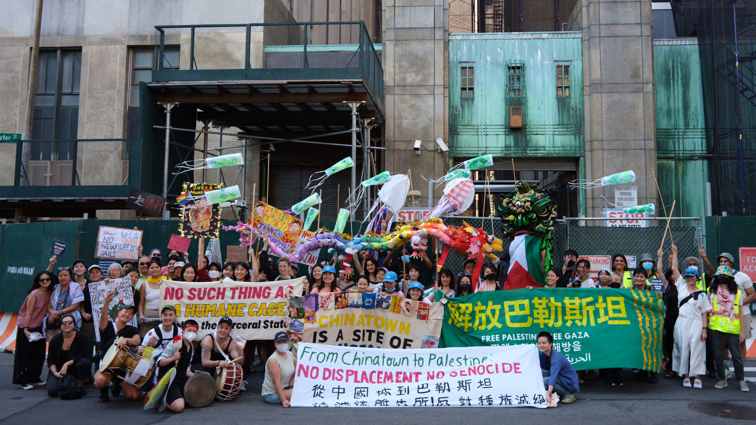 A diverse group of people is gathered in front of a construction site in an urban area, holding various colorful banners and signs. The banners contain messages...