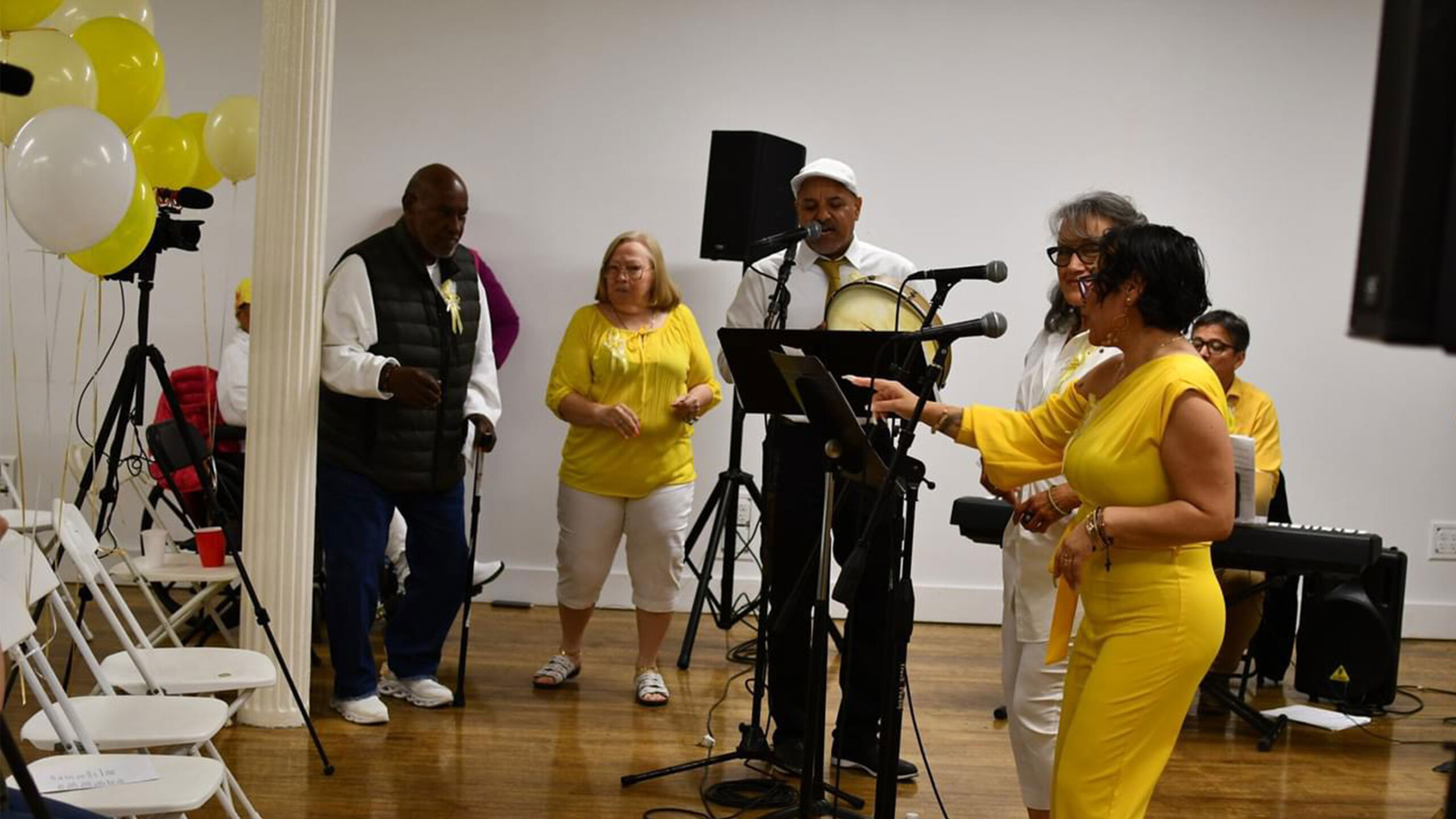 A lively group of people engaging in a musical gathering indoors. A woman in a bright yellow dress stands in the foreground while holding a microphone and addressing...