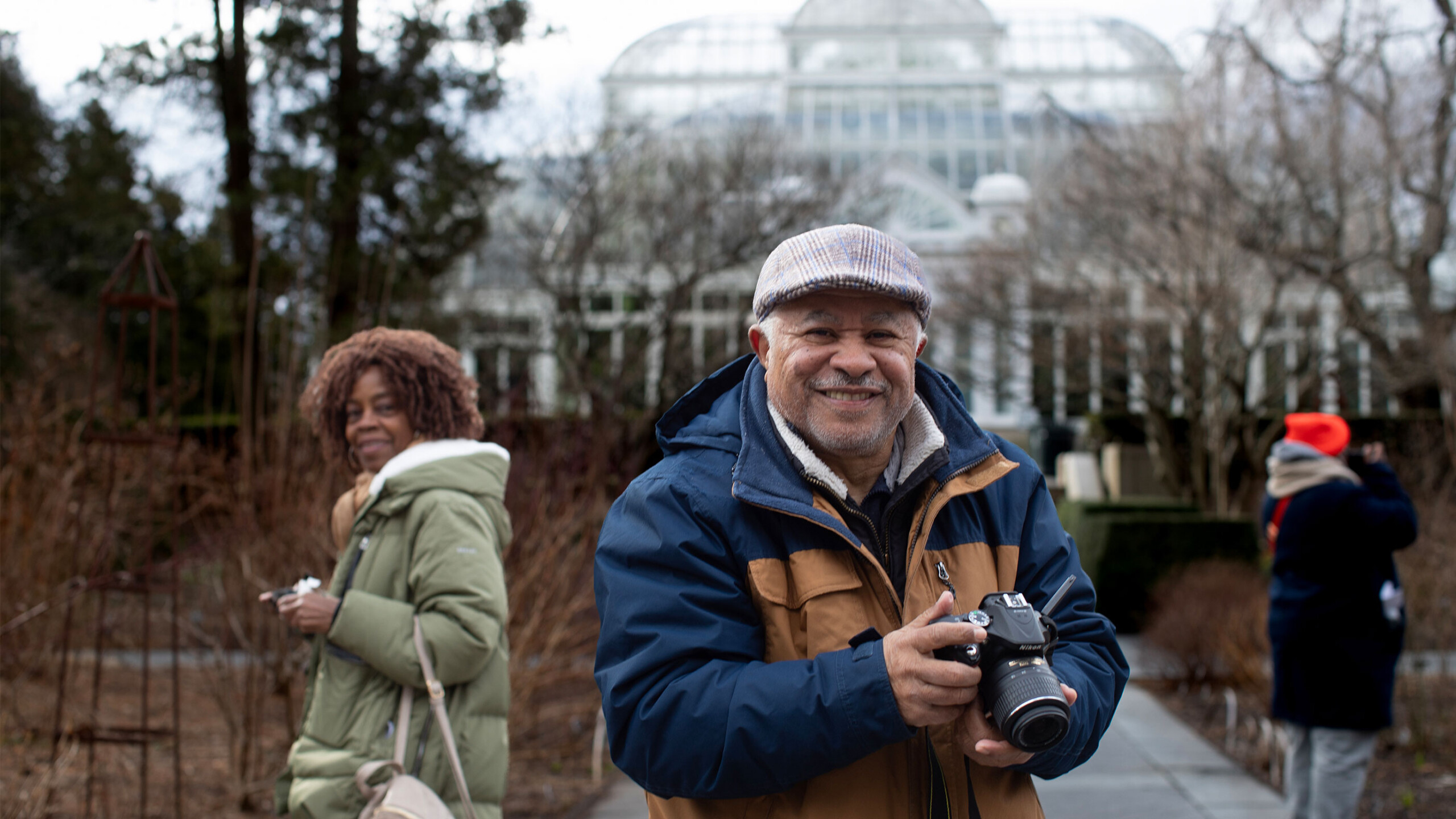A person holding a camera with a glasshouse in the background and another individual on the side.
