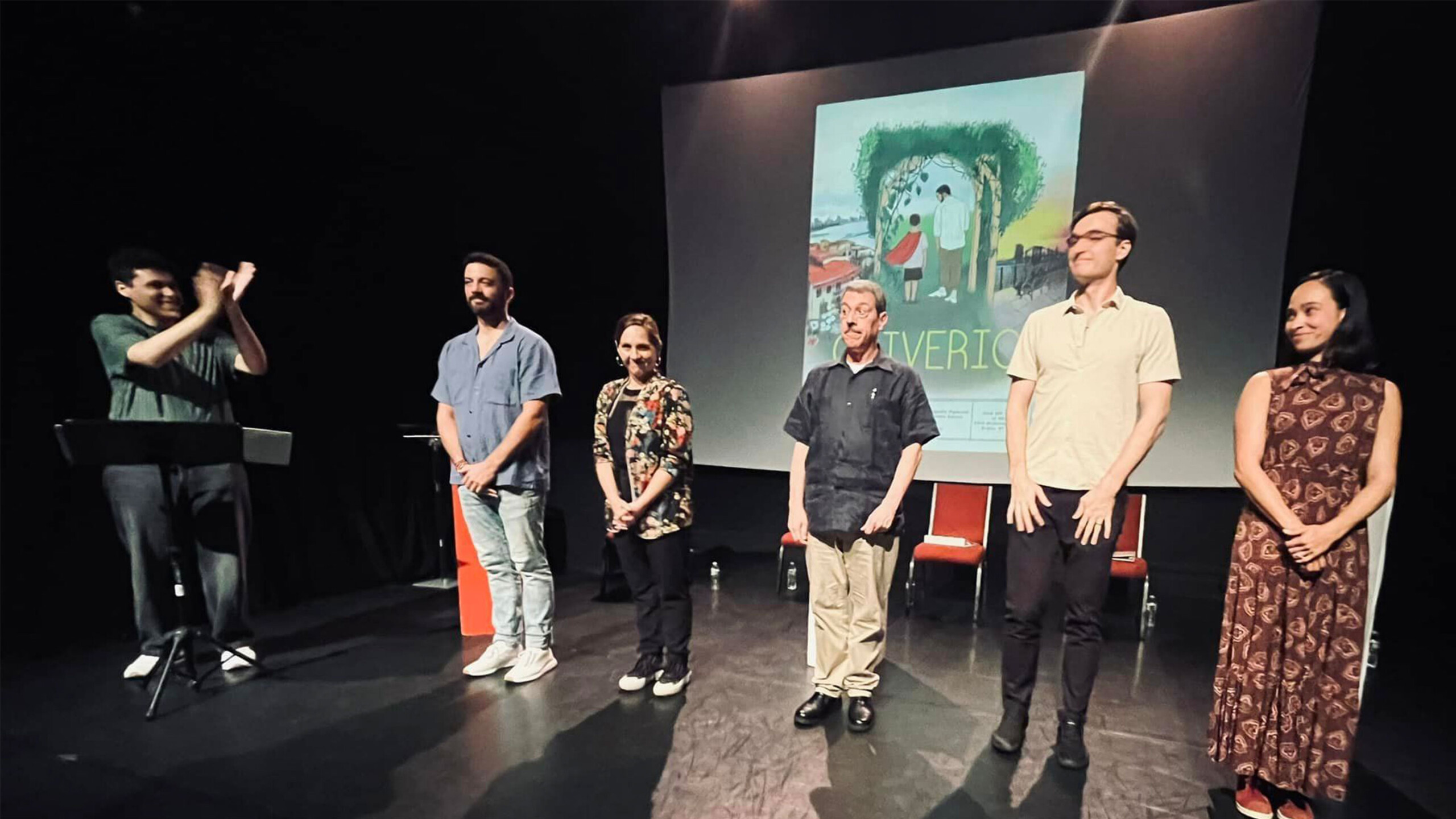 A group of six people stand on a stage, facing the audience. They are in a semi-circle under warm lighting, and all appear to be involved in some kind of performance...