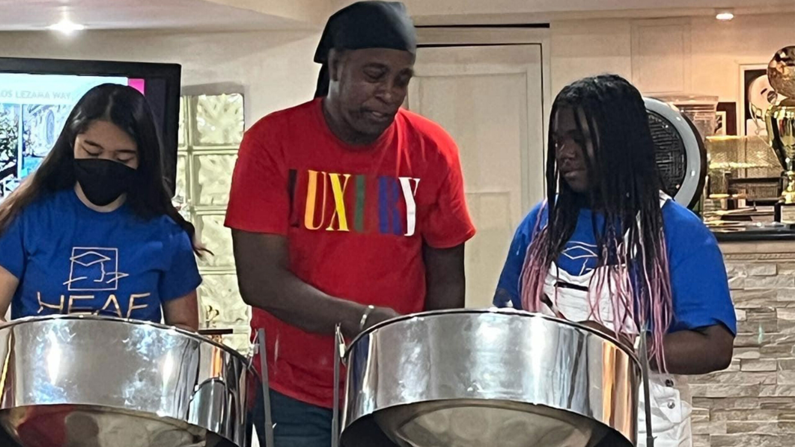 Three individuals are engaged in playing steel drums in an indoor setting. The person in the middle, wearing a red t-shirt and a black head covering, is assisting...