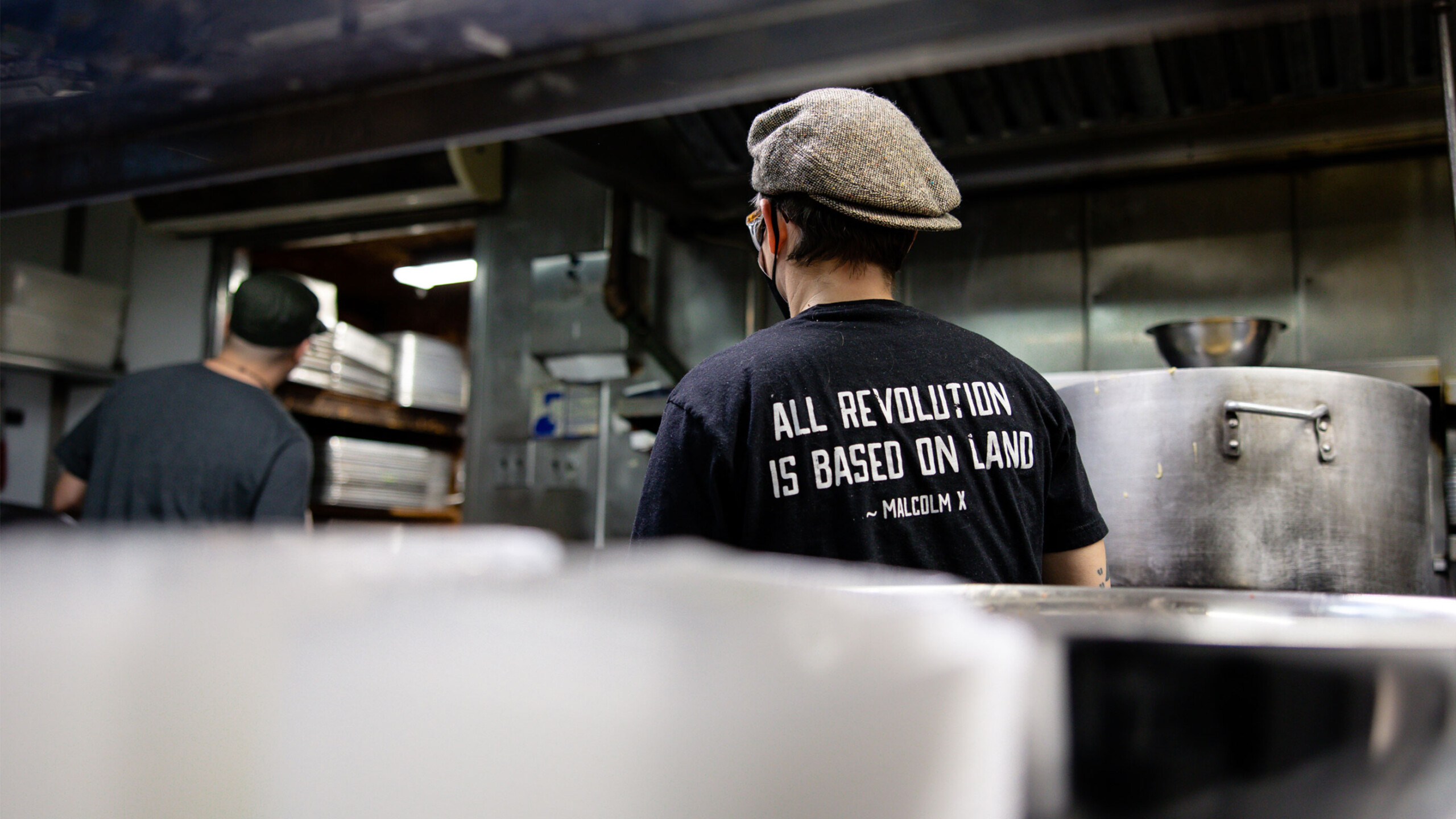 Two people working in a kitchen setting, with backs turned to the camera. The focus is on one person's black t-shirt, which has a quote from Malcolm X printed on...
