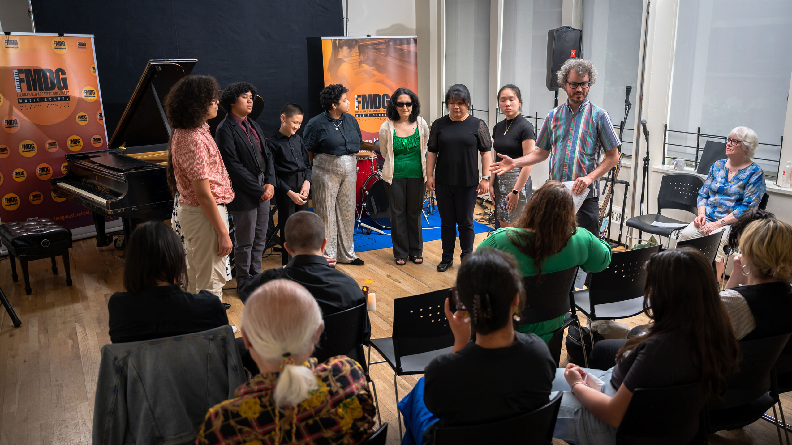 A group of people is gathered in a room, likely engaged in a musical event or rehearsal. The setting includes musical instruments such as a piano, and the decor...
