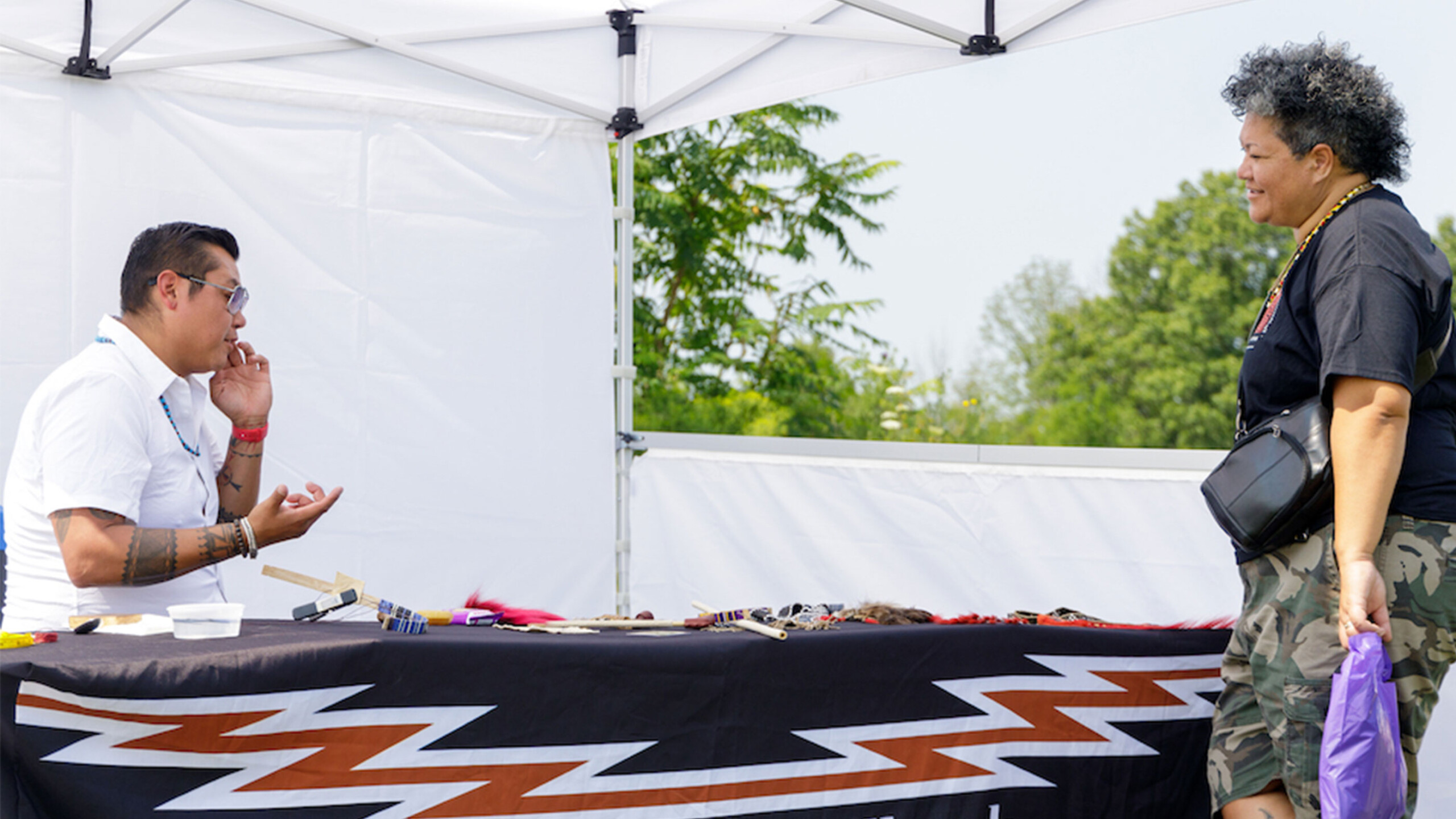 A man and a woman engage in conversation at an outdoor market stall. The man, seated and wearing a short-sleeved shirt and glasses, is speaking with the woman....