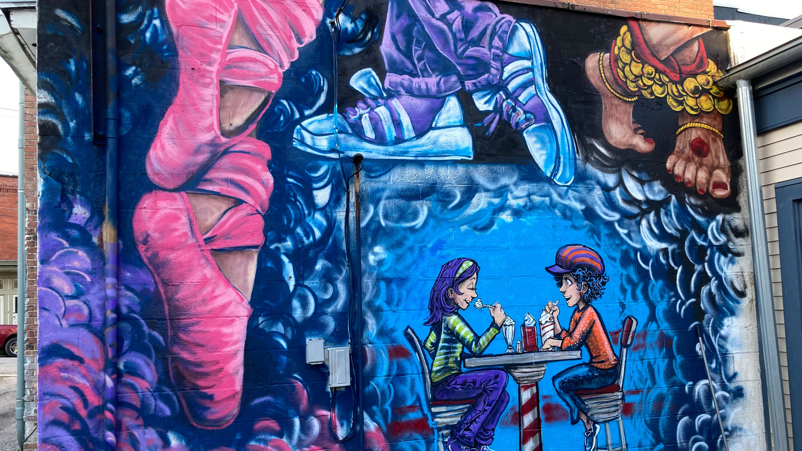 A colorful mural of two people at a table with drinks on a building's wall, under oversized dangling feet.