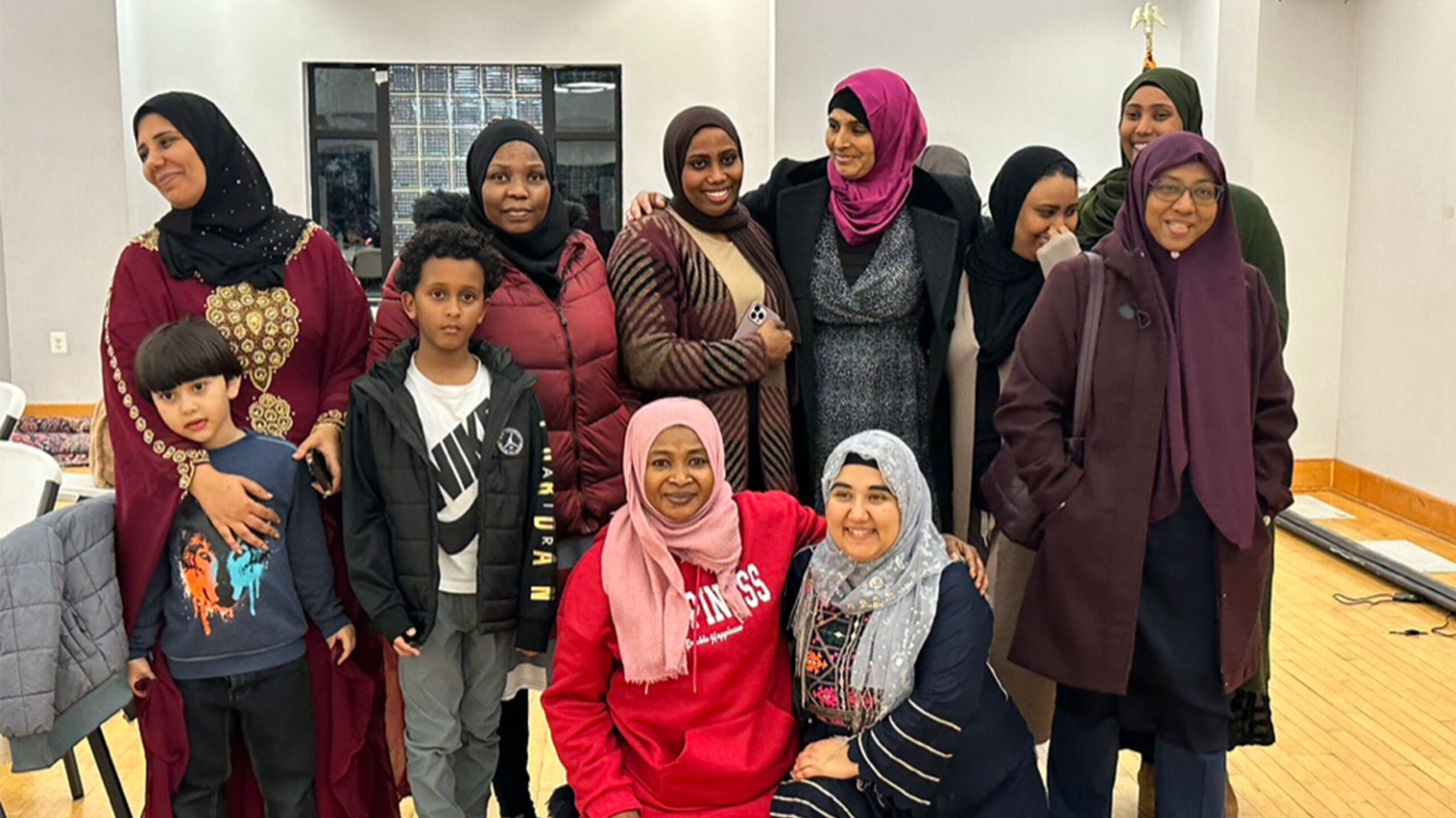 A diverse group of individuals gathers indoors, featuring nine women and two children. The women are dressed in various styles of hijabs and modest clothing with...