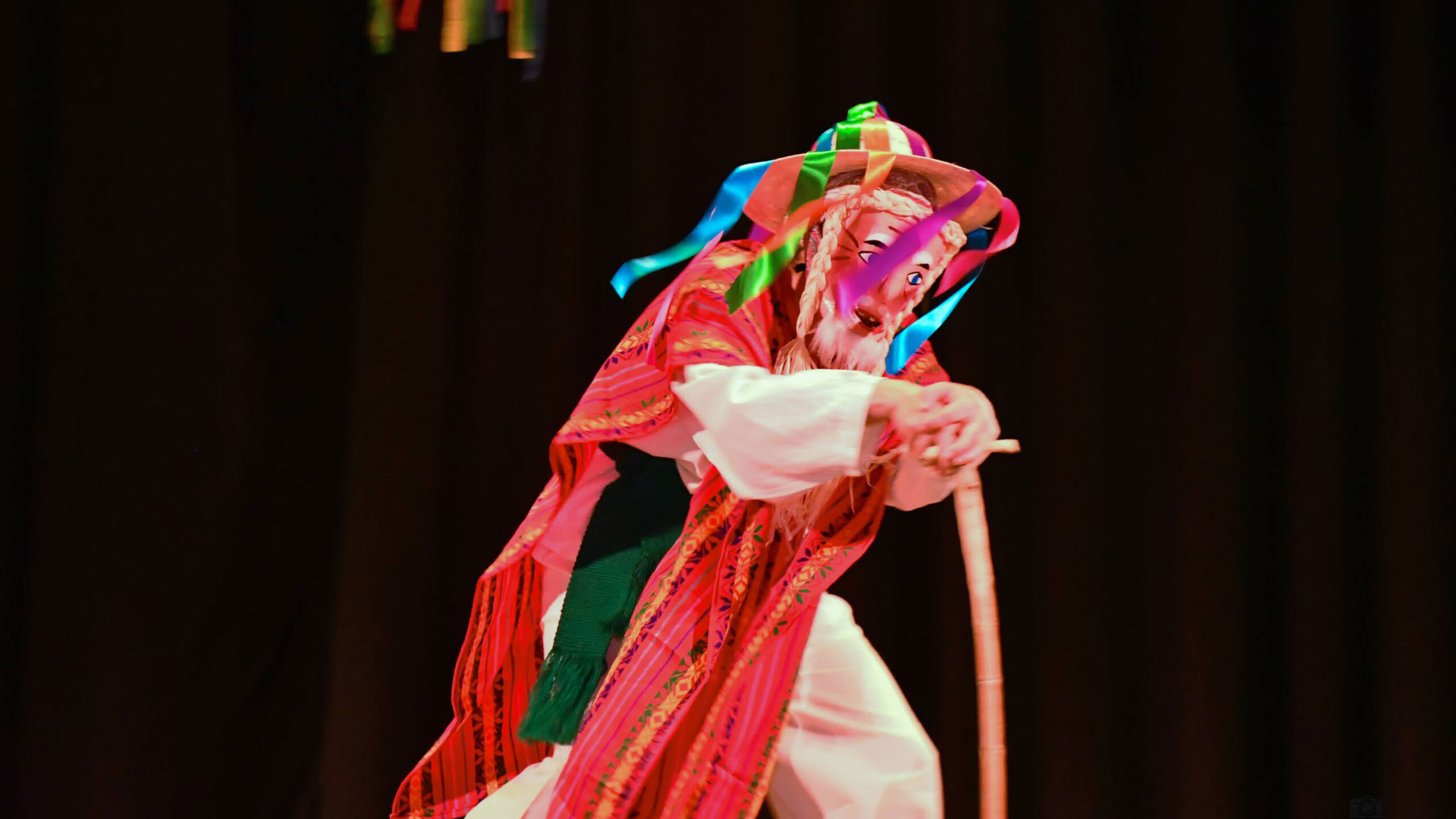 A performer dressed in a vibrant traditional costume dances on stage. The outfit includes a colorful striped poncho and a wide-brimmed hat adorned with brightly...