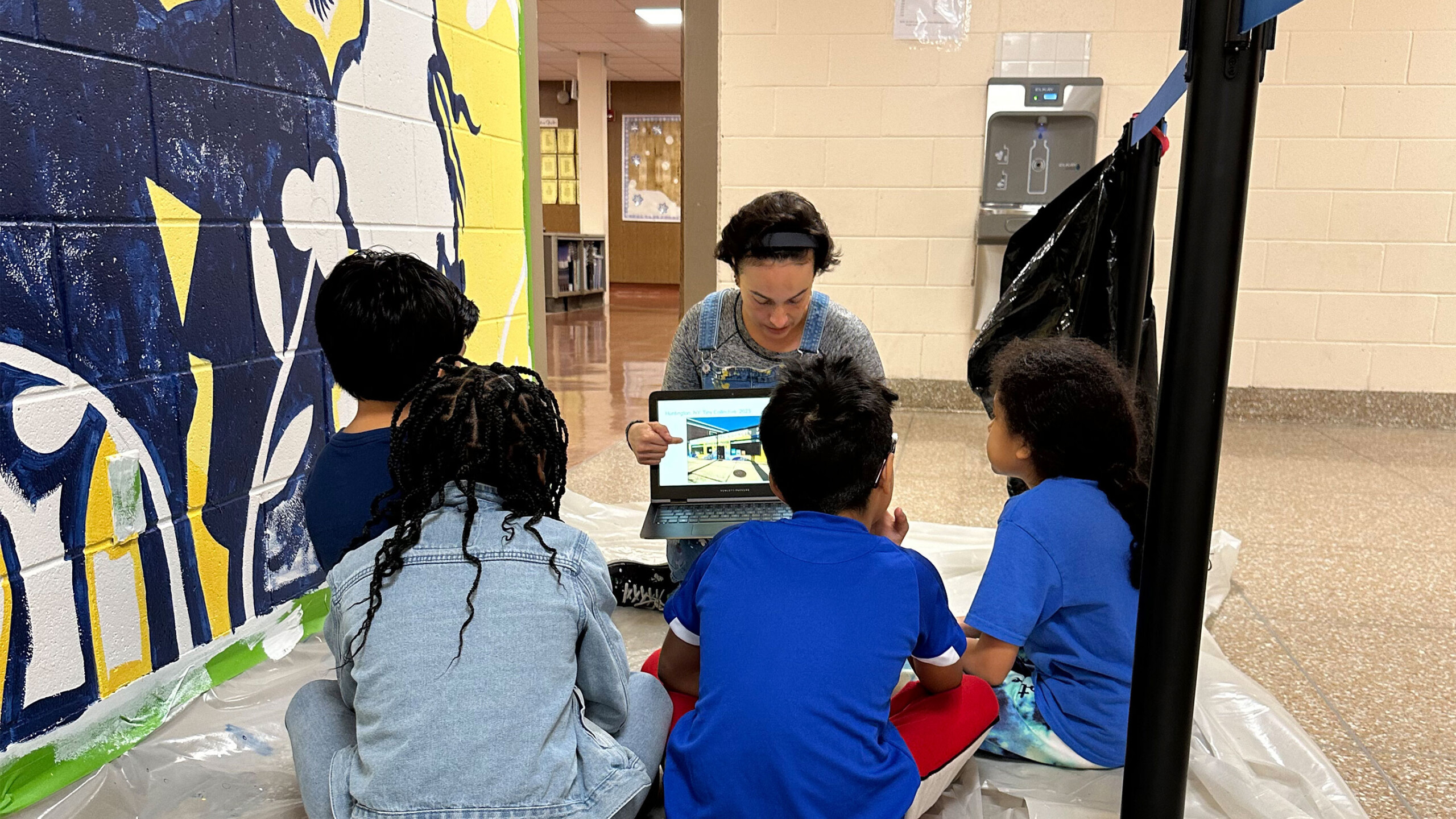 A group of four young children, dressed in casual attire, sit on a protective sheet on the floor, engaging with an adult who is showing them something on a screen....