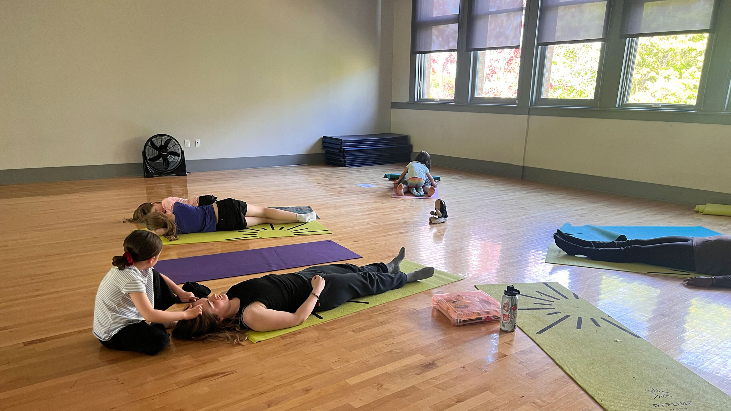 A spacious room with wooden floors accommodates a yoga class where participants are lying on colorful mats. One person is receiving a gentle head massage from an...