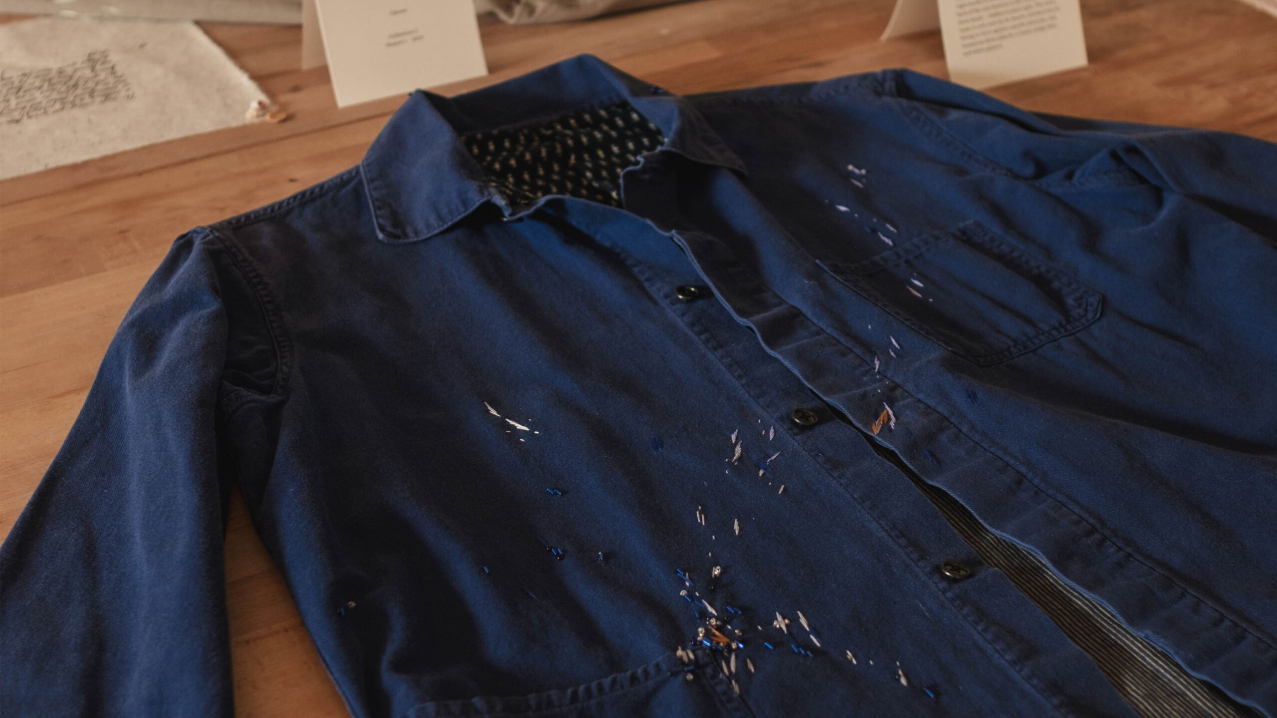A blue collared work shirt is laid flat on a wooden surface, possibly a tabletop. The shirt features several paint splatters, indicating it might have been used...