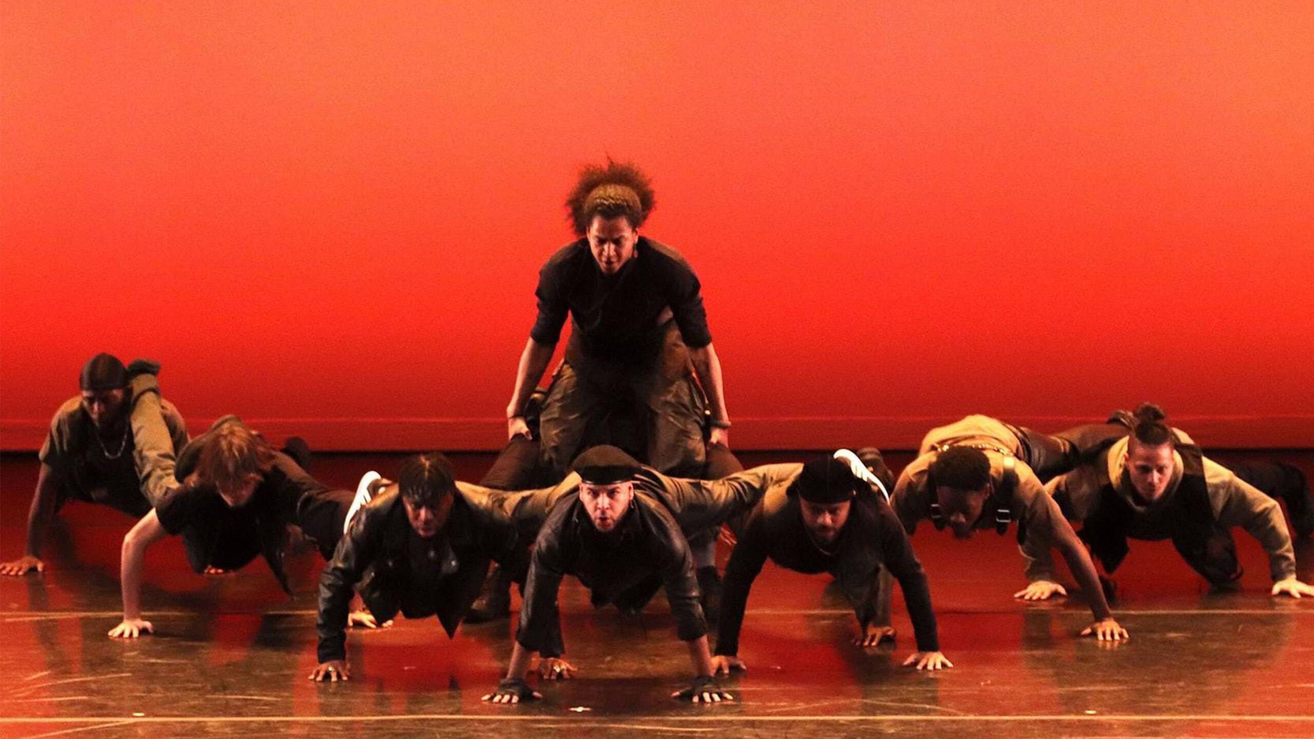A group of dancers performs an energetic routine on stage against a vibrant red-orange background. The formation consists of several dancers supporting one individual...