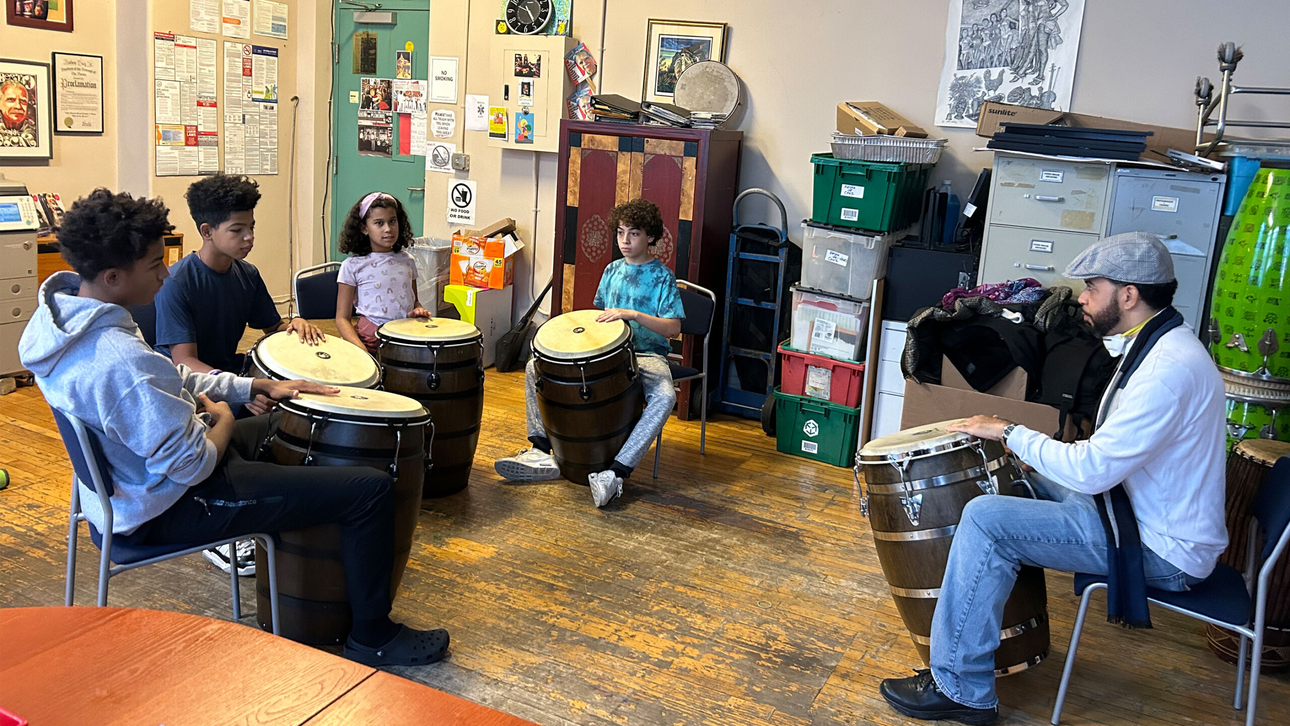 A group of four young students sitting in a semicircle, each with a conga drum in front of them, attentively watching an instructor or teacher who is seated and...