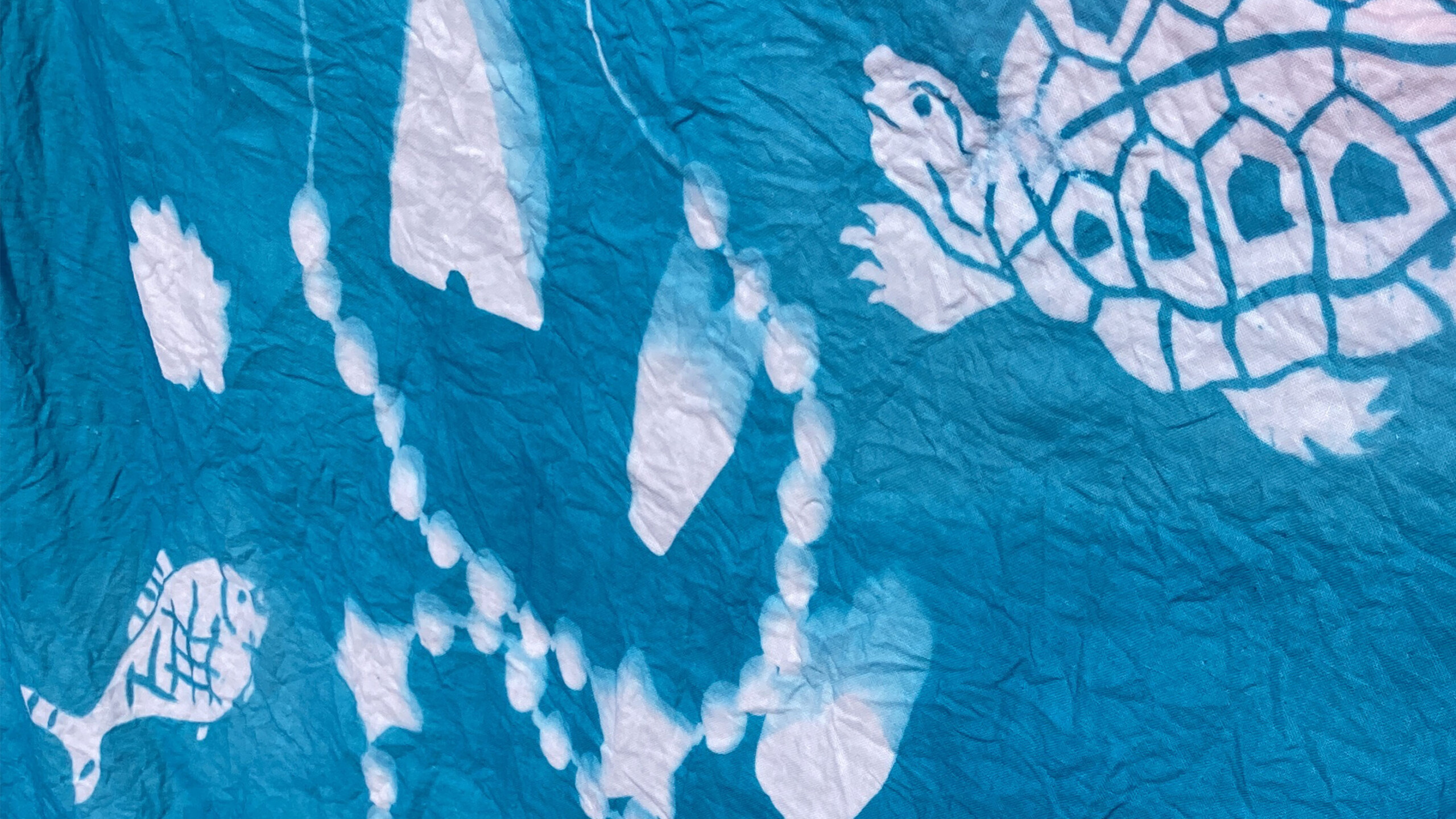 A crumpled blue fabric featuring a white batik design of marine life, including a large turtle and a small fish. The turtle is shown with a detailed shell pattern...