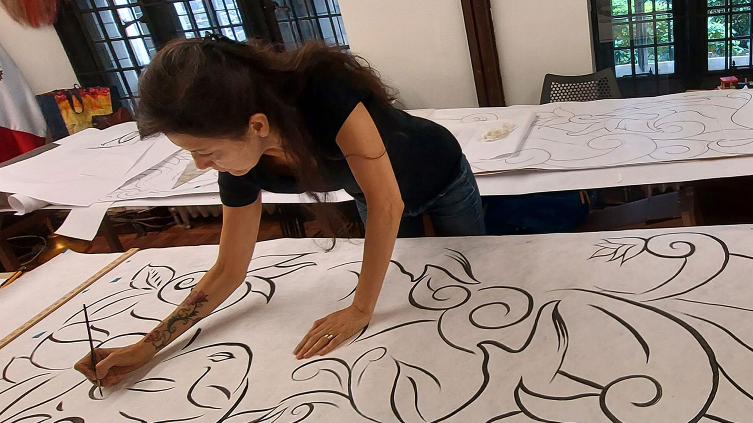 A female artist leans over a large canvas, intently focused as she paints intricate patterns with bold black lines. Her right hand guides the brush smoothly while...